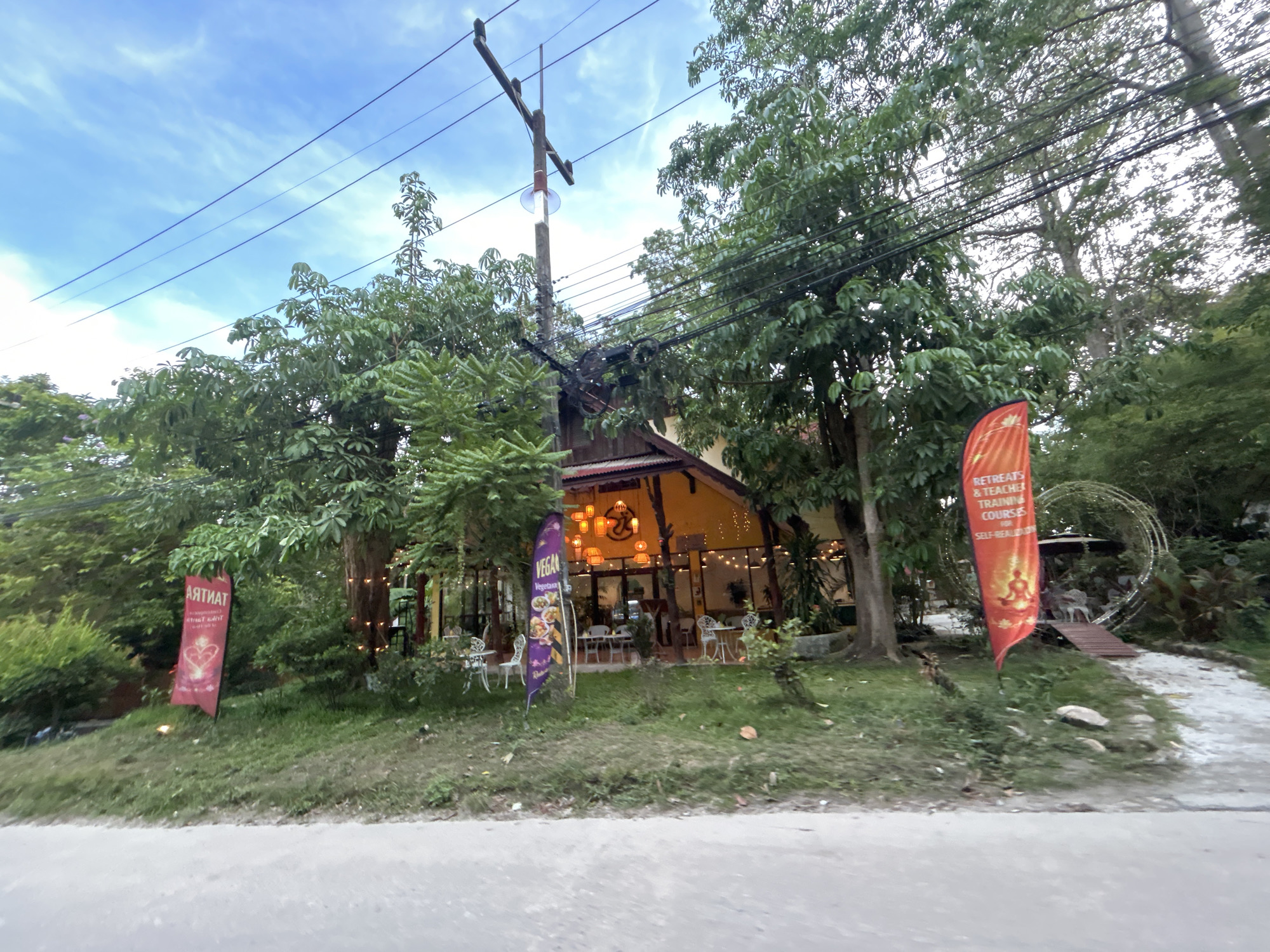 Captured from ground level in Baan Sri Thanu, Thailand, this exterior shot shows a restaurant with a wooden roof and glass front. The restaurant is nestled among trees and greenery. The street in front is slightly out of focus. Signage includes a banner promoting "Tantra" on the left and another indicating "Retreats & Teacher Training Courses" on the right. A "Vegan" banner is also visible near the building. String lights are hung inside the restaurant. Tables and chairs are set up inside and on the small front patio. An electric pole and wires are overhead. The sky is light blue with faint clouds.