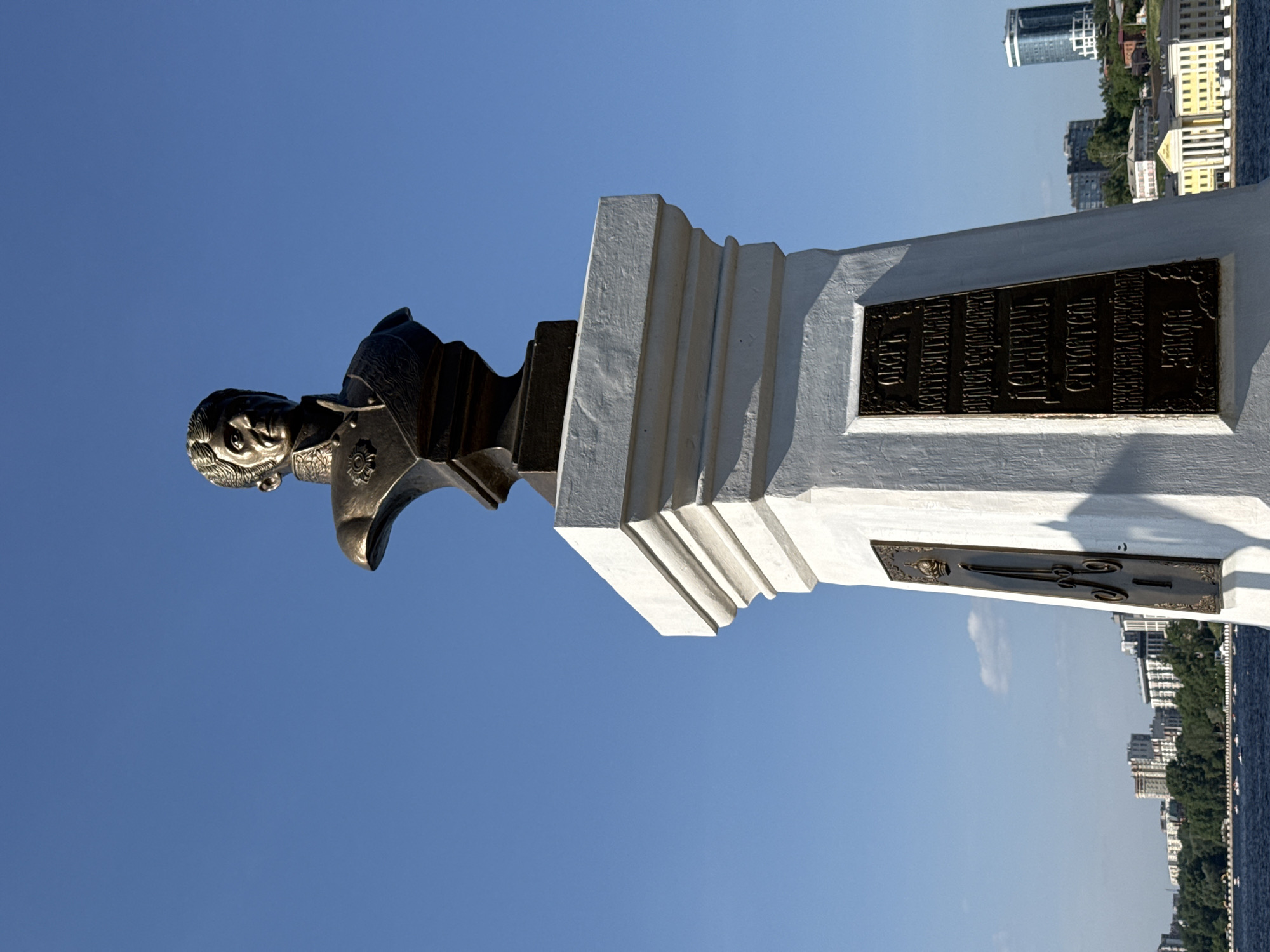 Here is a description of the image:
The image depicts a monument against a blue sky, likely located in Izhevsk, Russia. The monument consists of a dark metal bust mounted on a white, tiered pedestal. The pedestal features two bronze plaques with text, presumably inscriptions or historical information. In the background, glimpses of a cityscape with buildings, trees, and a body of water (likely a river or lake) are visible. The image is oriented sideways.