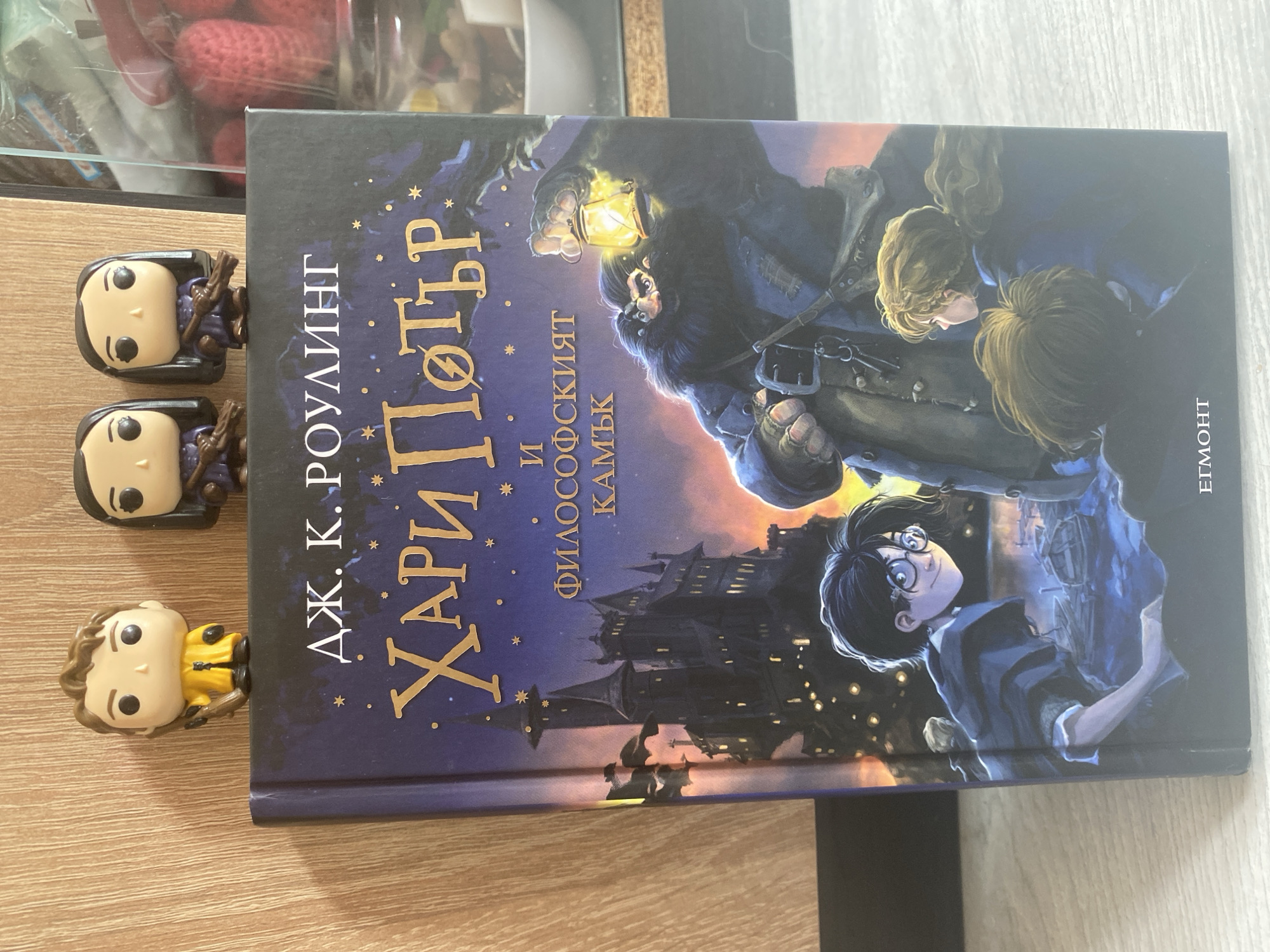 The image shows a close-up of the front cover of a Bulgarian edition of "Harry Potter and the Philosopher's Stone". The cover features an illustration of Hogwarts castle at night, with Hagrid holding a lantern and Harry Potter and other children in the foreground. Three Funko Pop figures of Harry Potter characters are perched on top of the book. The book is placed on a light-colored wooden surface, possibly a table or shelf. In the background, out of focus, is a clear plastic container filled with red, crochet-like objects. The lighting suggests an indoor setting, and the overall presentation indicates a personal collection or display. There is no discernible information in the image that would allow for pinpointing a specific location within Blagoevgrad, Bulgaria; the scene is generic and could be anywhere.