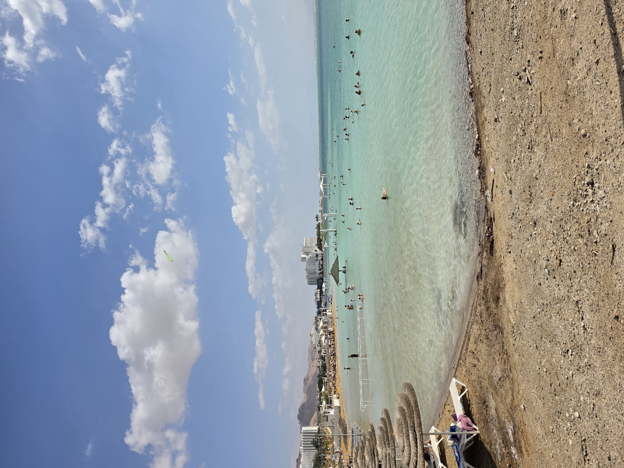 The image shows a beach scene in the city of Tamar Regional Council, Israel. The Dead Sea occupies the right half of the image with its characteristically turquoise water, where several people are floating. The shore has a sandy texture. Along the shoreline, there are sun umbrellas and lounge chairs. A white structure extends into the water. In the background, buildings and mountains are visible. On the left side of the image is a blue sky with white clouds. A green kite is also visible in the sky.