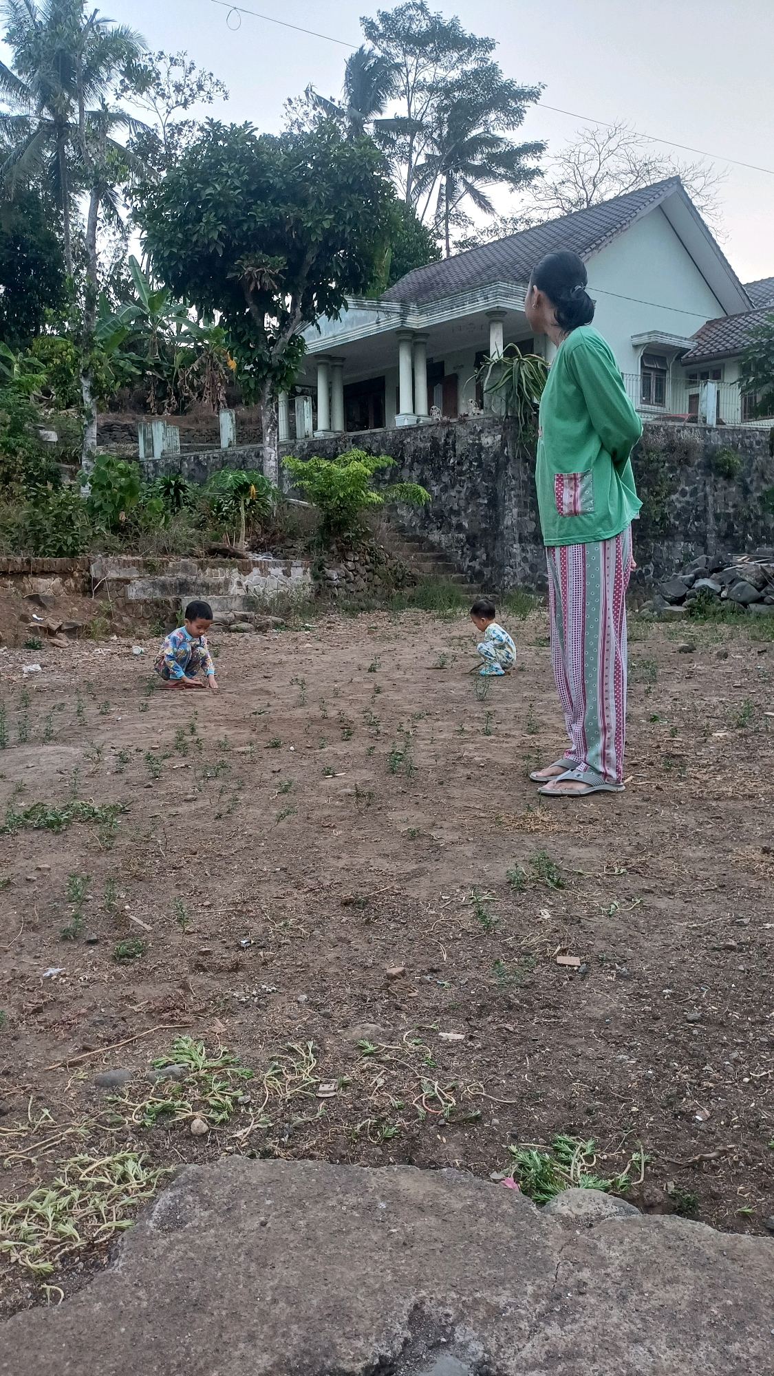 In Wonokerso, Indonesia, a woman in a green top and striped pants stands in a dirt yard, looking towards a house. Two young children are also in the yard, one on the left appearing to play with dirt and the other on the right crouching. The house, with a white exterior and a tiled roof, is visible in the background, along with trees and a stone wall. The yard is mostly bare earth with sparse green vegetation.