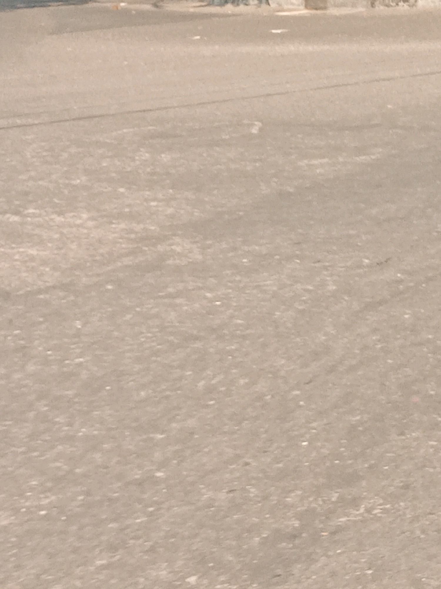 The image shows a close-up, ground-level view of an asphalt road in the city of Yogyakarta, Indonesia. The road surface is textured with small pebbles and appears dry. A faint, thin, dark line, possibly a shadow or a crack, runs diagonally across the upper middle portion of the frame. In the distance, at the very top of the image, a lighter-colored curb or pavement edge is visible, with a few small, indistinct objects that could be litter. The overall scene depicts a mundane urban environment without any prominent features or activity.