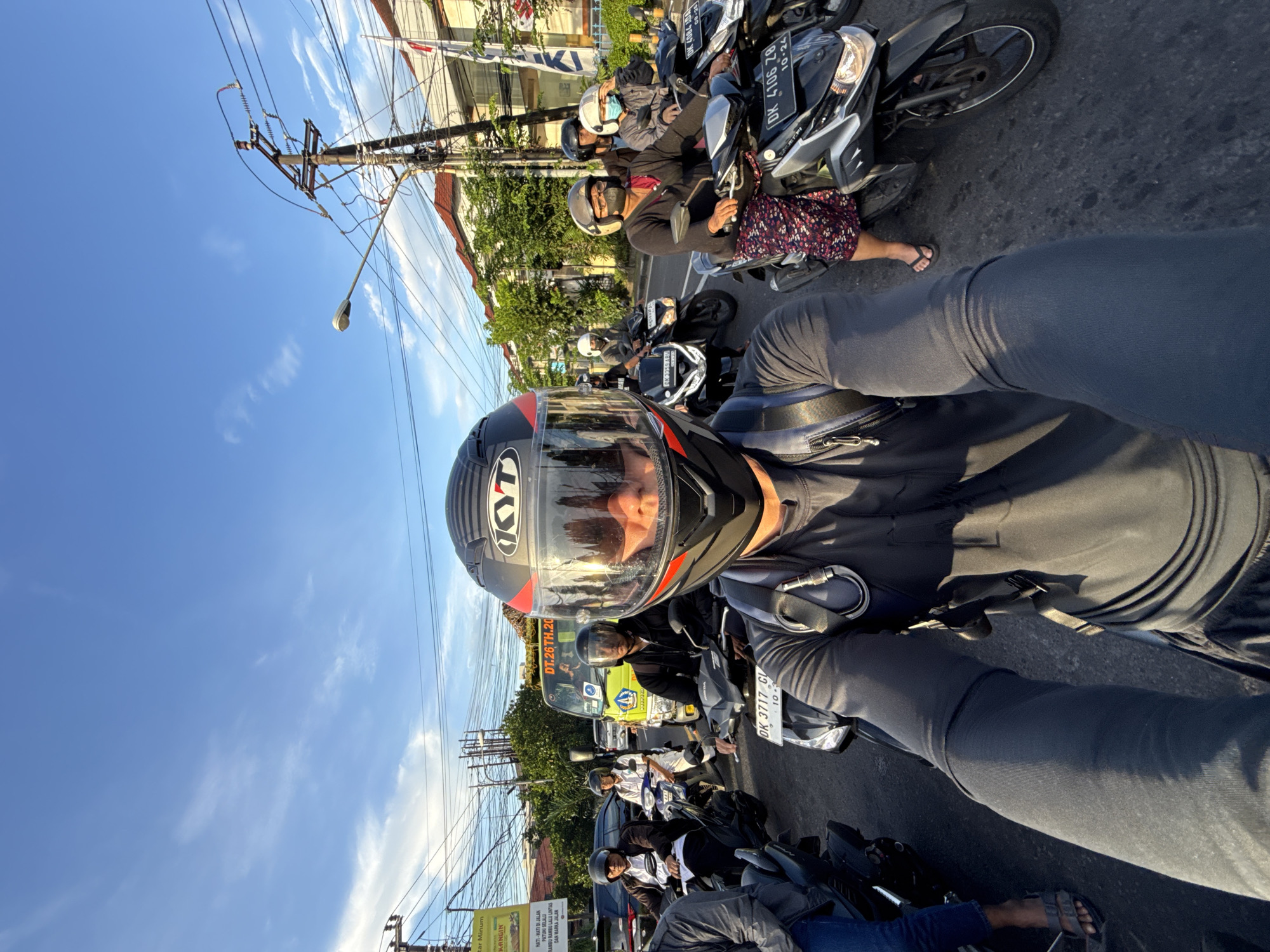 The image captures a first-person perspective selfie of an individual wearing a black "KYT" helmet, seated on a scooter, likely in traffic or stopped on a street in Denpasar, Indonesia. The main subject's face is visible through the helmet's clear visor.
The scene is bustling with multiple other people on scooters, many also wearing helmets and some with face masks, indicating a common mode of transport in the region. Directly to the right of the main subject, a person in a patterned skirt and flip-flops is visible on another scooter. Further down the street, more scooters and a yellow truck or bus can be seen.
The activity appears to be navigating or waiting in street traffic. The setting is an urban street with asphalt pavement, lined by buildings, green trees, and numerous utility poles with complex wiring. The bright, clear blue sky with wispy clouds suggests a sunny daytime, possibly morning or late afternoon, given the sunlight and shadows.
Notable details include the widespread use of scooters for transportation and the street infrastructure.
Visible text includes:
* "KYT" on the main subject's helmet.
* "HONDA" on one of the scooters.
* License plates starting with "DK", common in Bali, Indonesia: "DK 3717 CI" (with "10-3"), "DK 4084 ADA" (with "08-20"), "DK 4106 ZB" (with "10-24"), and "DK 3359 ABM".
* A green and yellow street sign in Indonesian, partially obscured, reads: "HATI-HATI DI JALAN" (Be careful on the road), "PATUHI SELALU RAMBU RAMBU LALU LINTAS" (Always obey traffic signs), and "DAN MARKA JALAN" (and road markings). Other words visible on signs are "tar Minum" and "KANGIN".
* "DT.26 TH.20" on the side of a vehicle in the background.