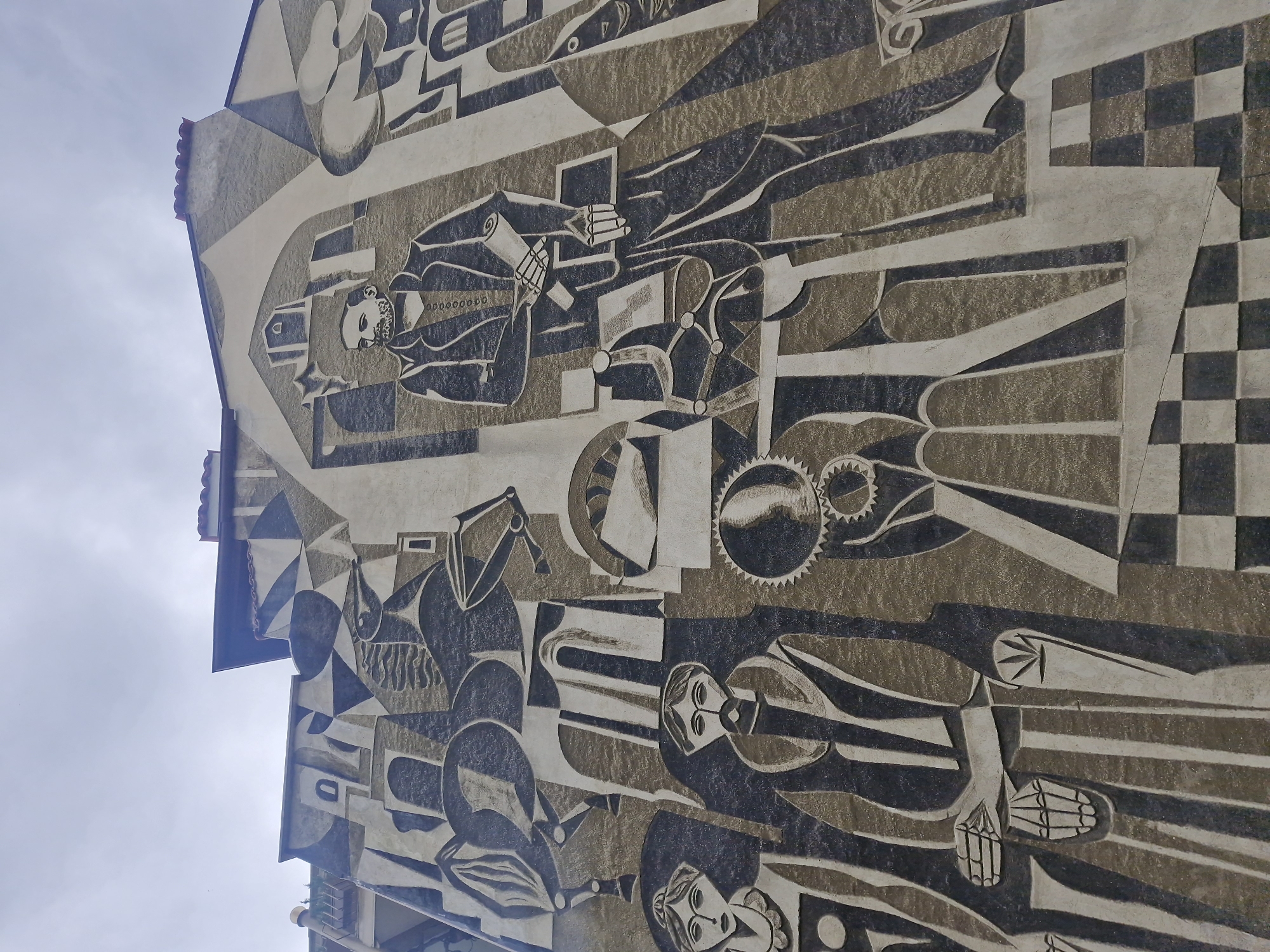 A low-angle shot captures a large, stylized mural on the side of a building in Plovdiv, Bulgaria. The mural features a complex arrangement of abstract figures and geometric shapes, rendered in a muted palette of gray, white, and black. The subjects depicted appear to be human figures in various poses and occupations, although the exact context is unclear. The artistic style suggests a Cubist or Constructivist influence. The sky is visible in the upper left corner, hinting at an outdoor setting. The building's architecture includes a sloping roofline, adding to the overall composition.