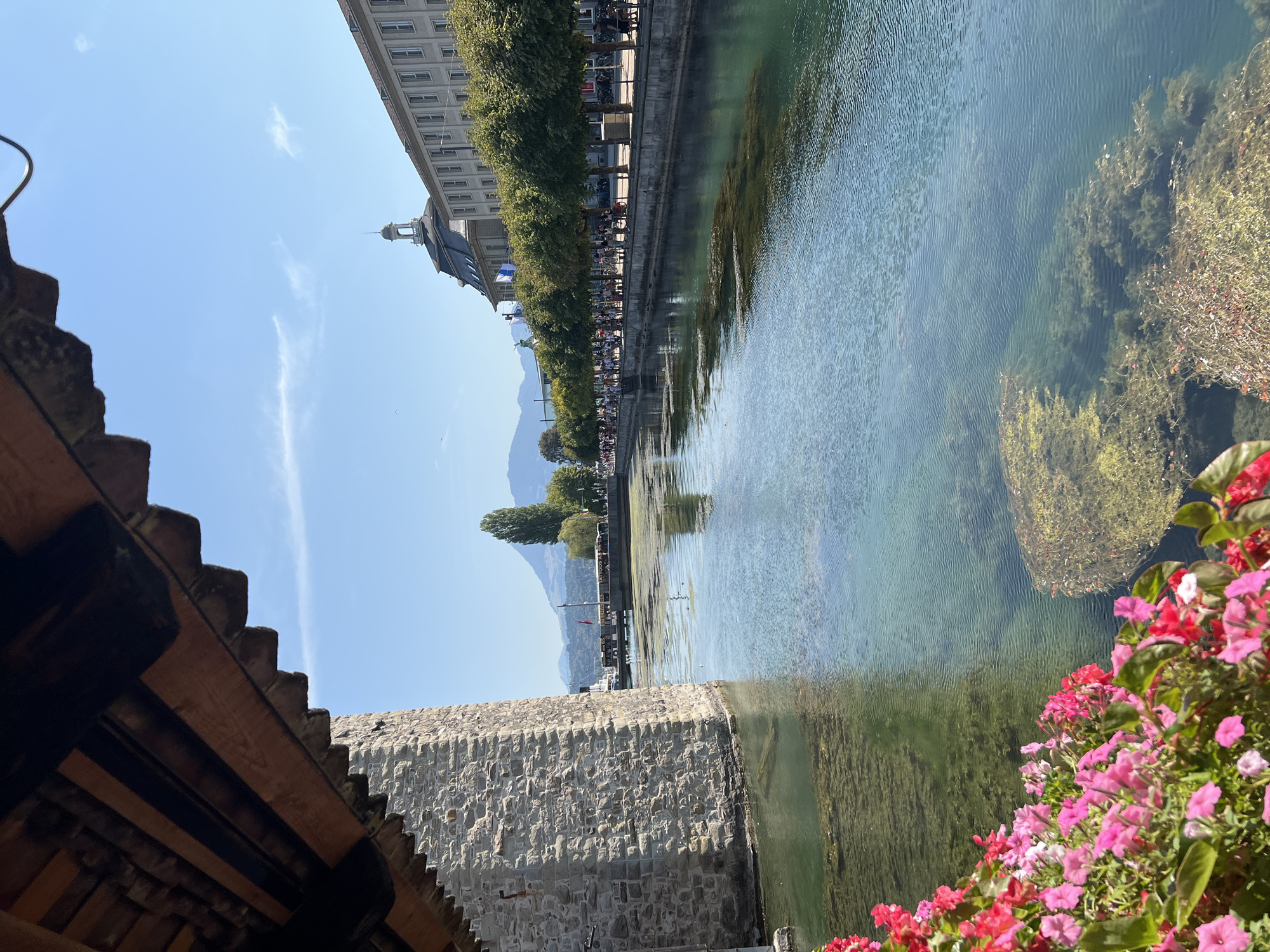 The image shows a clear, blue sky over a canal in Kriens, Switzerland. A stone tower is on the left, with a wooden roof overhead. On the right, buildings line the canal, with trees and people walking along the sidewalk. The water is clear, with some submerged vegetation visible. There are some pink flowers in the foreground on the left.