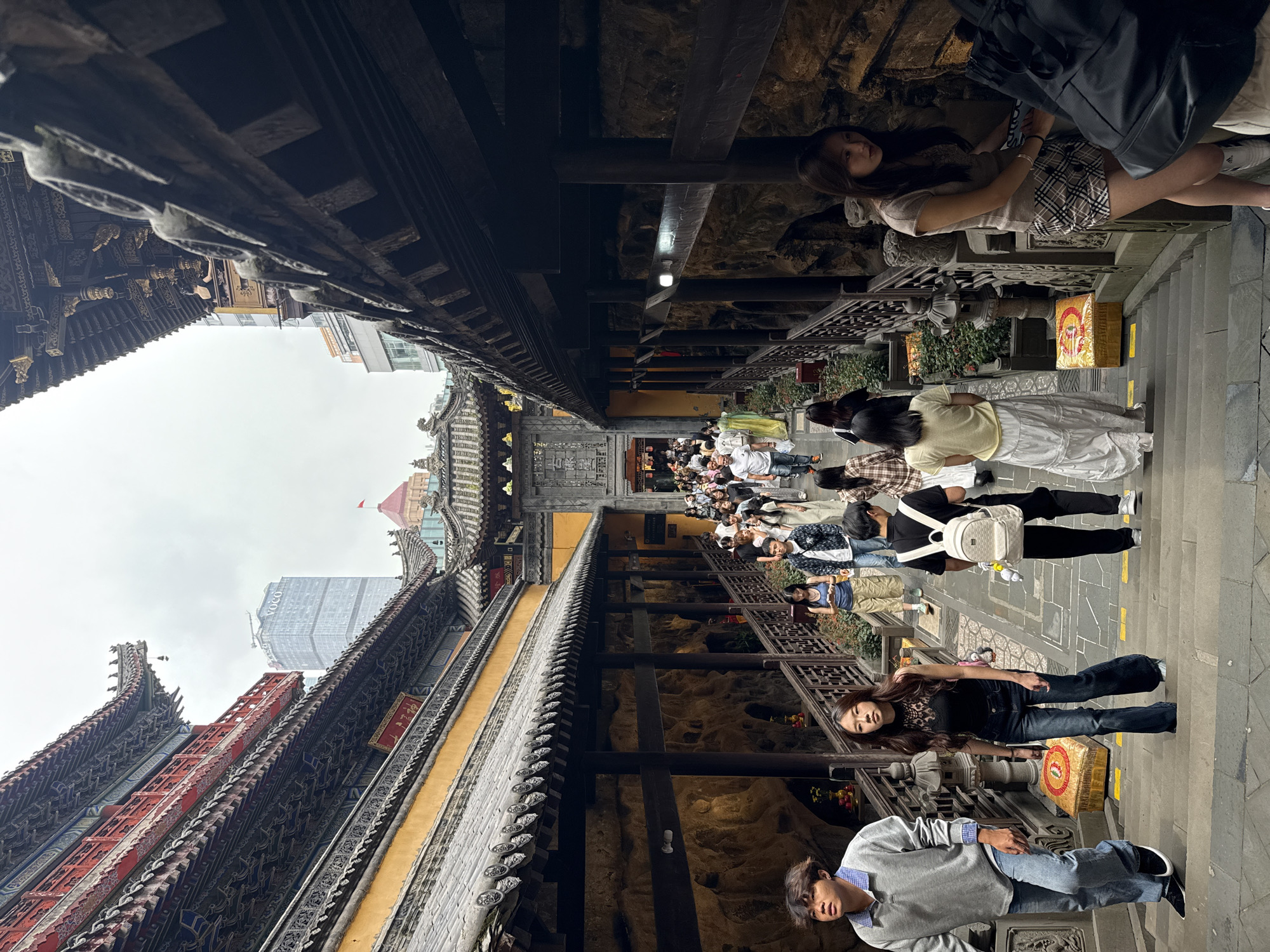 Captured in the Yuzhong District of China, the photograph presents a crowded stone pathway leading towards a traditional Chinese temple or monument. The buildings are adorned with intricate, weathered architectural elements and feature roofs with upturned eaves, indicative of Chinese design. A modern high-rise building looms in the background, juxtaposing the ancient structures with contemporary urban development.
Numerous people are present, some standing and observing, while others are walking through the area. Some individuals are seen interacting with their mobile phones. The overall ambiance suggests a bustling tourist spot or public area where history meets the modern world.