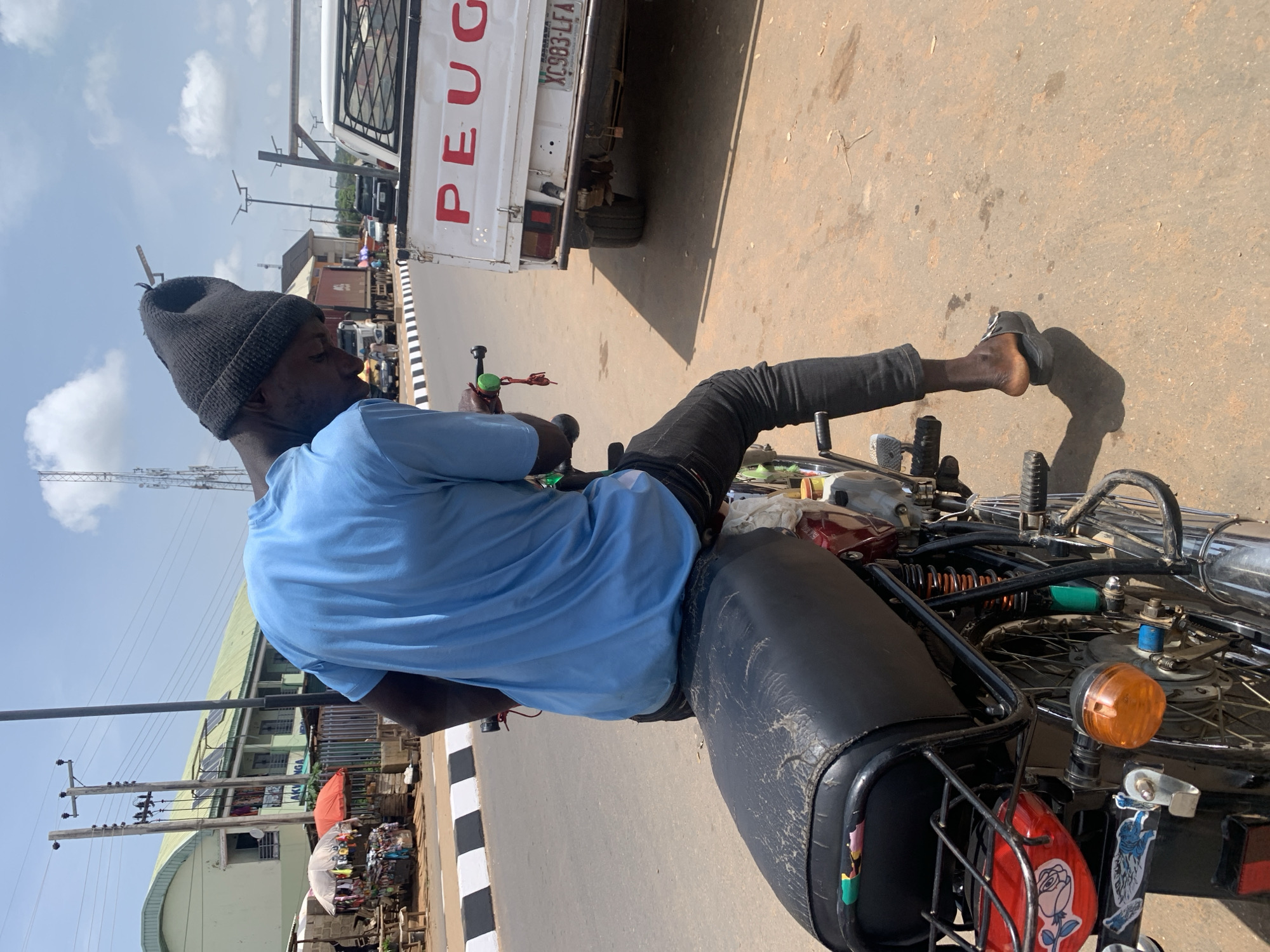 In Akwanga, Nigeria, a young man wearing a light blue t-shirt and black beanie sits on a black motorcycle. He is facing away from the camera, looking down at the motorcycle's handlebars. The motorcycle is parked on an asphalt road with a white and black striped curb. In the background, there are shops and buildings, including a green-roofed building with solar panels and a white Peugeot pickup truck. The sky is partly cloudy.