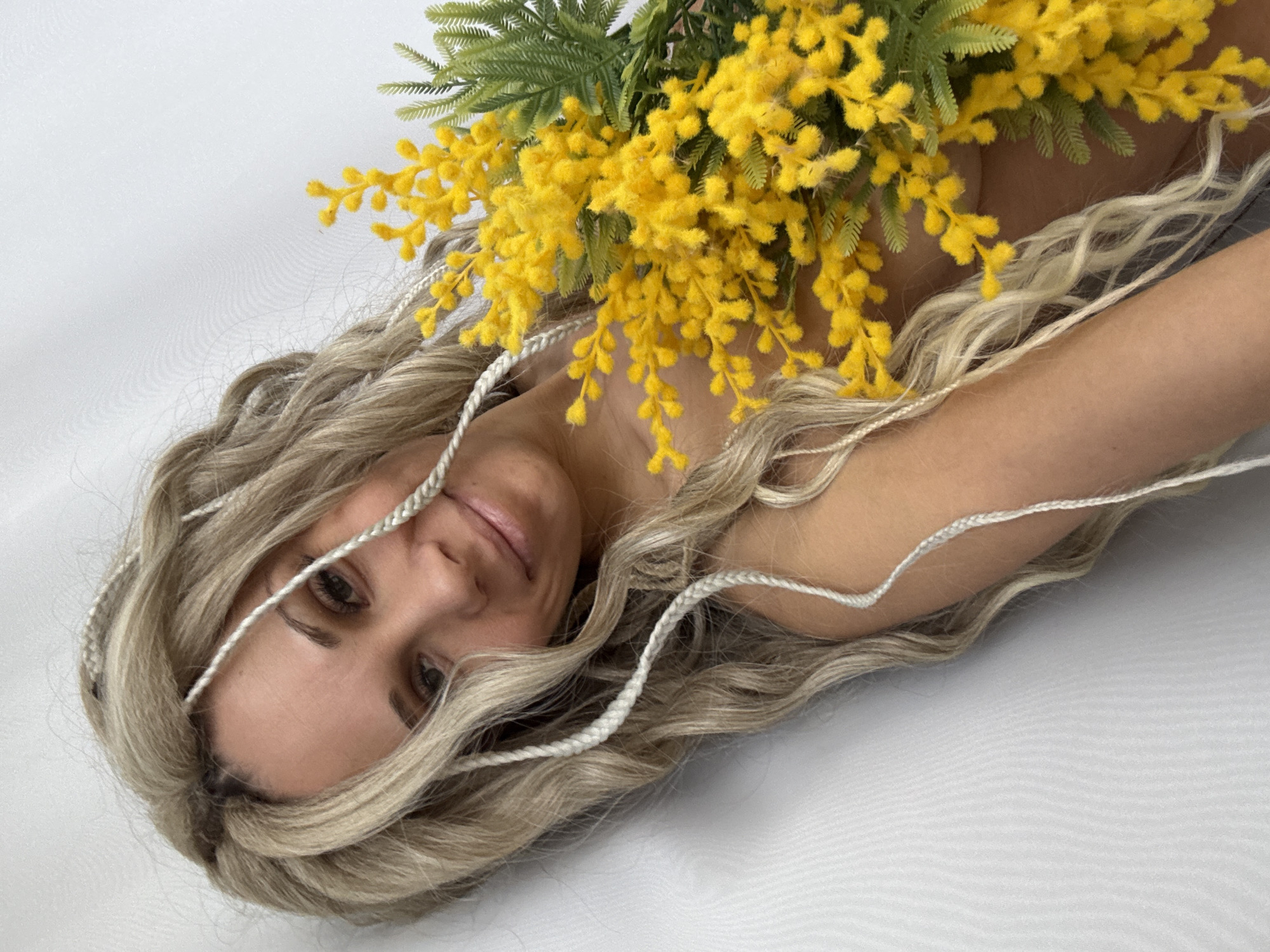 A woman with blond, wavy hair is lying on a white surface. Her hair is styled with white braids. She is holding a bouquet of yellow mimosa flowers. Her face is visible, and she is looking towards the viewer with a slight smile. The background is plain white.
