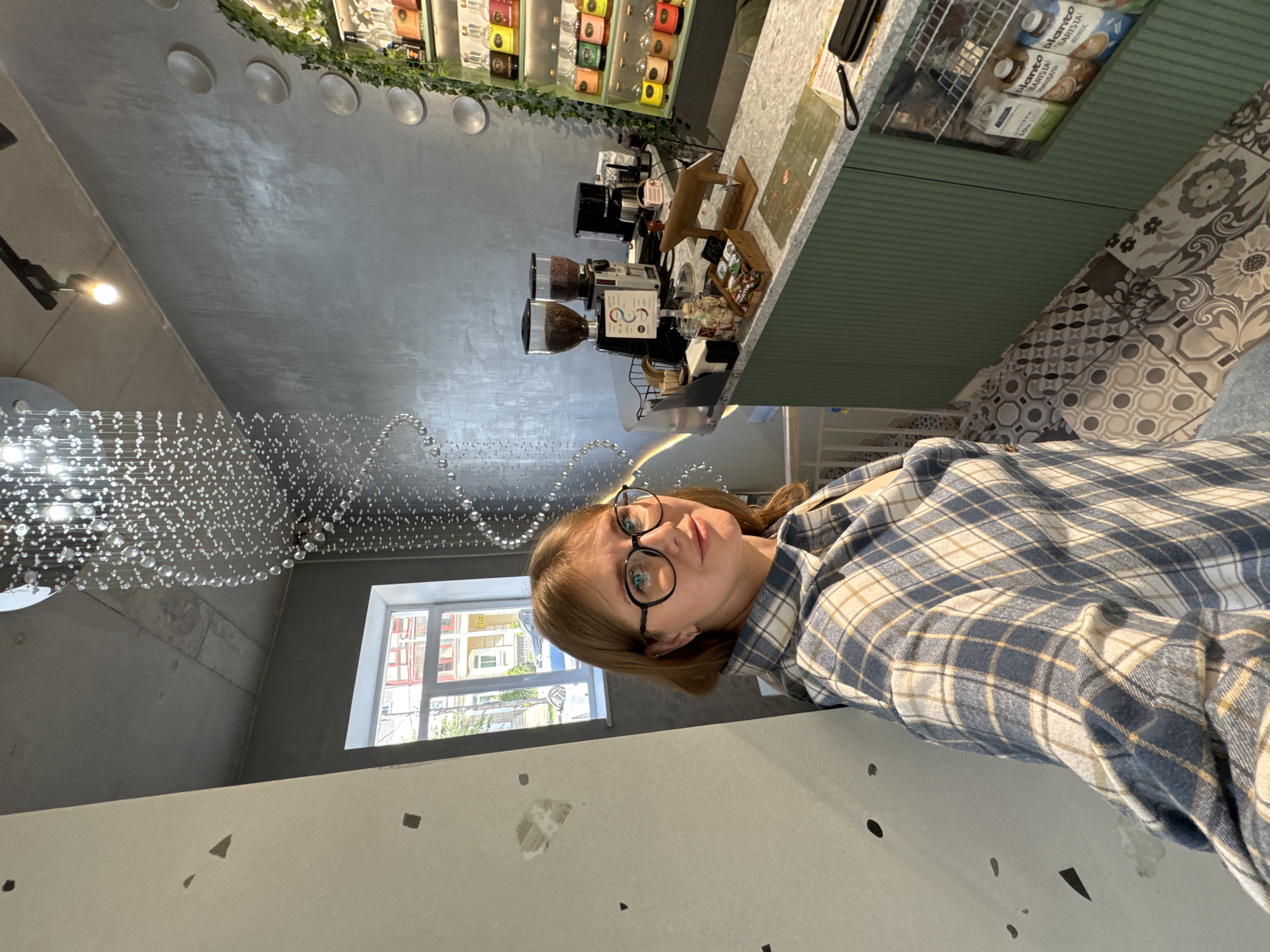 This is a medium shot, selfie-style photo taken indoors. A young woman with shoulder-length brown hair and glasses is in the foreground, looking at the camera. She is wearing a blue and white plaid shirt. She is standing in what appears to be a cafe or coffee shop, with a counter to her right. The counter has coffee grinders, a coffee maker, and bottles of syrup displayed on shelves behind it. The cafe has modern decor with a concrete ceiling, a decorative light fixture hanging from the ceiling, and patterned floor tiles. The background shows a window with a view of buildings outside, suggesting an urban location. The overall lighting is bright, likely from natural light and interior lighting. The exact location within the city of Perm, Russia, is not discernible from the image alone.