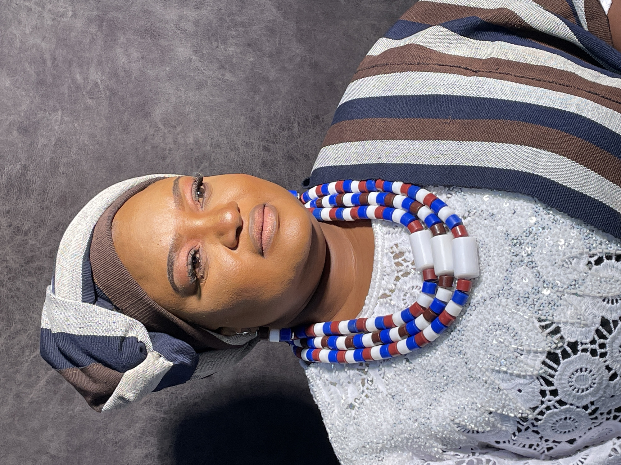 A woman is pictured against a dark, textured backdrop, possibly a studio setting. She's wearing traditional Nigerian attire, including a striped headwrap in shades of brown, navy, and white, and a white, beaded lace top. Around her neck is a thick, three-strand necklace of red, white, and blue beads, punctuated by larger white cylindrical beads. The lighting highlights her facial features and attire. There is no indication from the image that it is in Barakin Akawo, Nigeria.