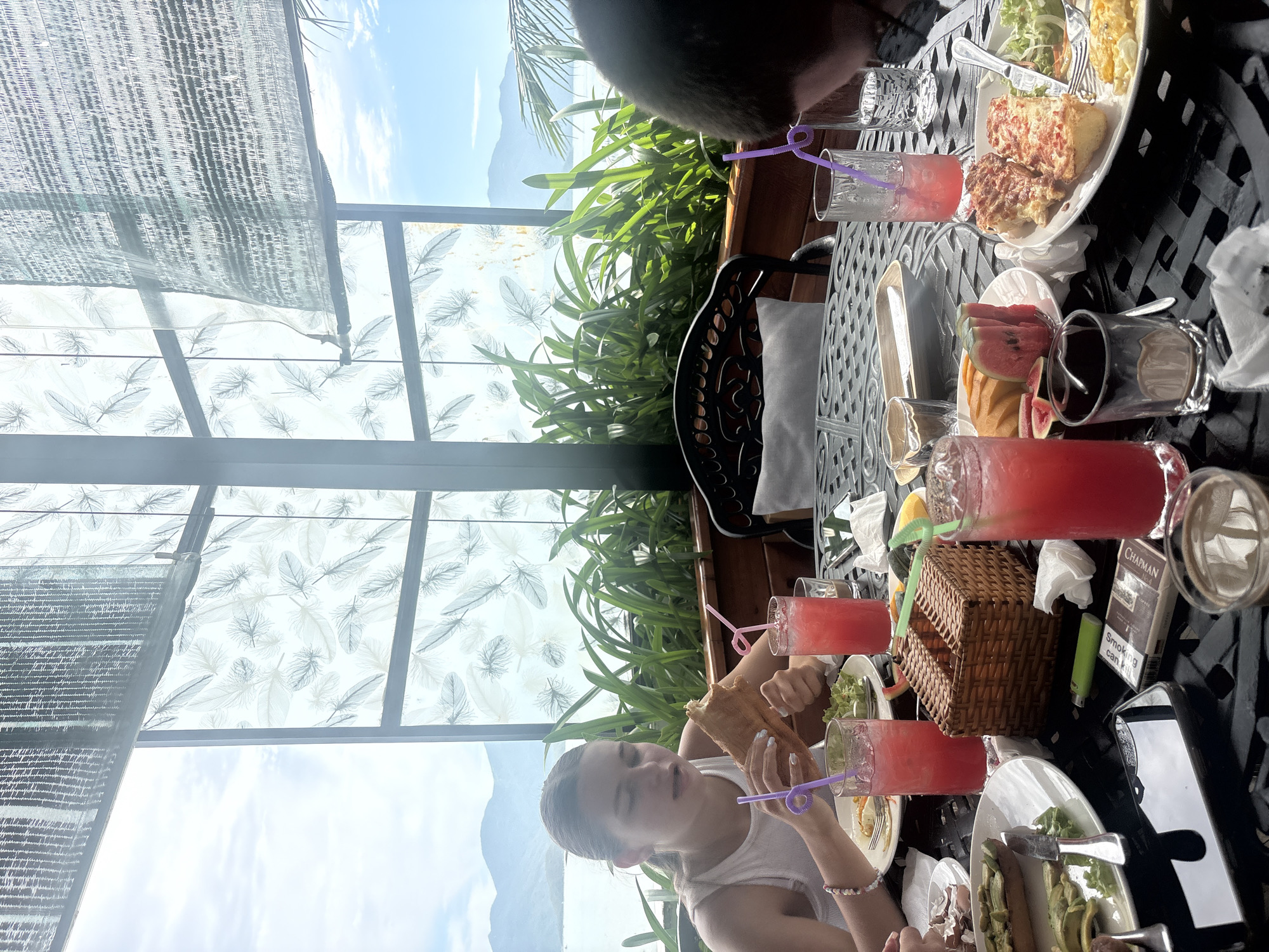 This is a medium shot of a group of people seated at a black, ornate metal table outdoors, likely at a restaurant or cafe in Bắc Nha Trang, Vietnam. The table is laden with food and drinks, suggesting a meal is in progress.
In the foreground, a young woman with brown hair pulled back is actively eating a large sandwich. She is wearing a white tank top and has a bracelet on her left wrist. In front of her are two glasses with pink beverages and purple straws. To her left, on the table, are plates with food, including what appears to be avocado toast and some type of cooked eggs or pastry. A cell phone, a lighter, and a pack of cigarettes are also visible on the table.
To the right of the frame, another person, partially visible from behind, has their head turned down towards their food and drink. They also have a pink beverage with a purple straw. Further back on the table, a small wicker basket is present, along with plates of food including sliced watermelon, cantaloupe, and another dish with what looks like baked cheese or pizza.
The background reveals a large window or glass enclosure with a decorative pattern of feathers. Beyond this, a body of water and distant mountains are visible under a bright, cloudy sky. Lush green plants are growing in planters behind the table, creating a semi-enclosed, scenic dining area. The overall atmosphere appears casual and relaxed, with the natural light suggesting it is daytime.