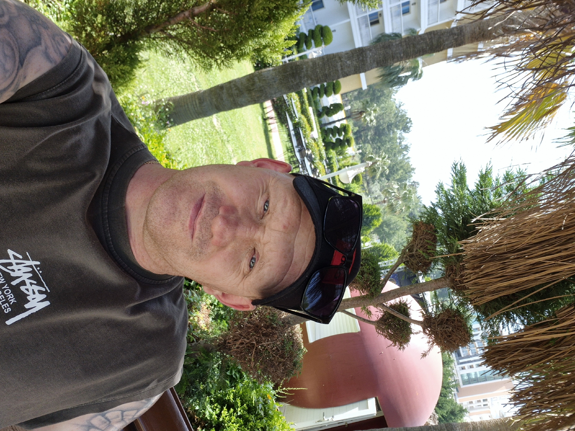 The image is a selfie of a Caucasian man with short, light-colored hair, a slight stubble, and sunglasses on his head. He is wearing a dark green t-shirt with white lettering on the left side that reads "STUSSY NEW YORK LOS ANGELES". Tattoos are visible on his left arm. He appears to be outdoors in a sunny location, likely in Alanya, Turkey.
In the background, there is a lush green garden with palm trees and meticulously trimmed bushes. A tall, multi-story building is visible in the distance. The image is rotated clockwise.