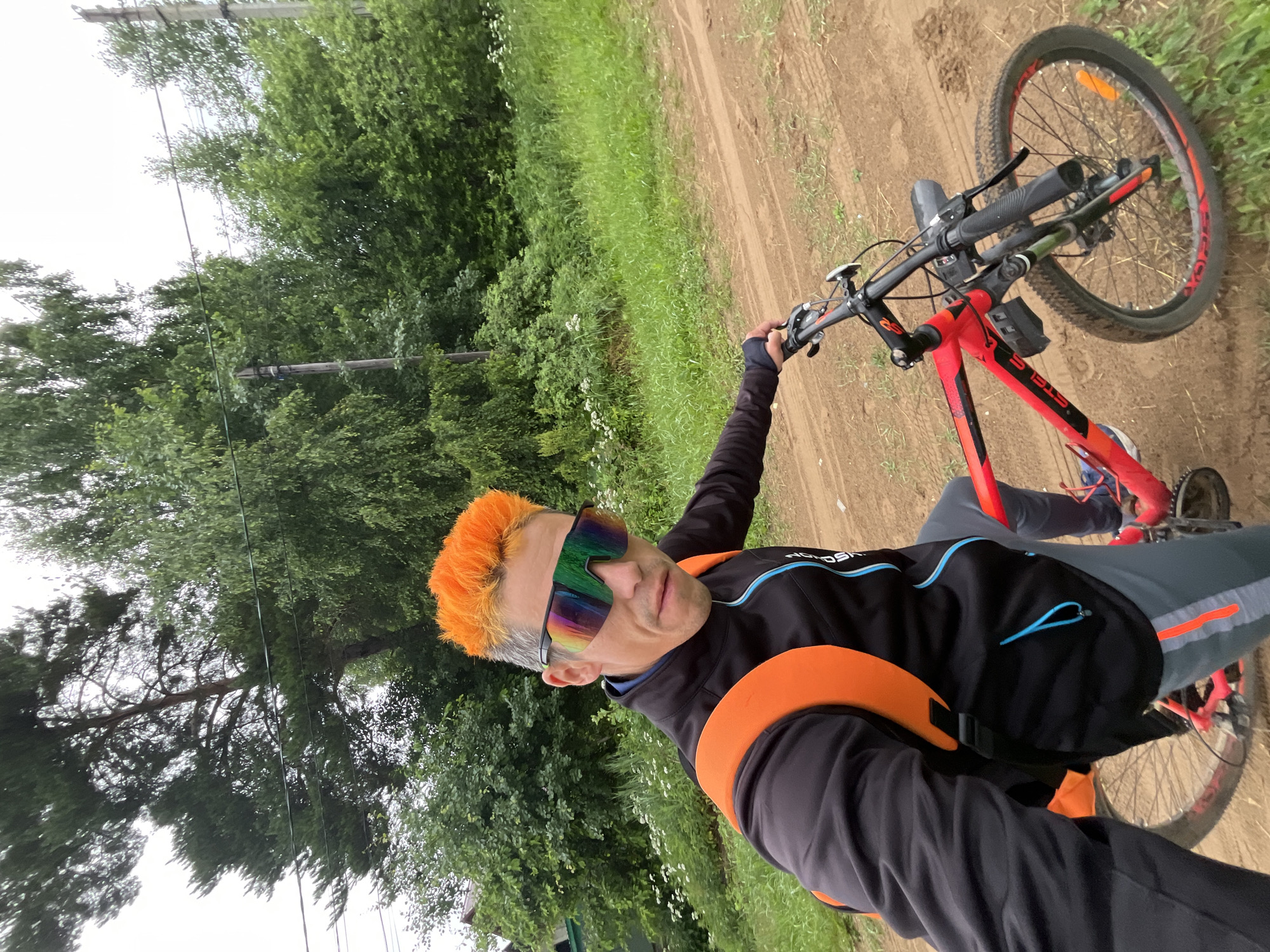 A man with bright orange hair is taking a selfie while riding a red bicycle on a dirt road. He is wearing a black jacket with orange and blue accents, grey pants with orange stripes, and large, multi-colored cycling sunglasses. An orange backpack is visible on his back. The background consists of lush green trees and foliage. The road appears to be in a rural or semi-rural area, possibly on the outskirts of Chaikovsky, Russia, given the presence of trees, vegetation, and a dirt path that could be a typical rural road. The overall lighting suggests an overcast or diffused daylight condition.