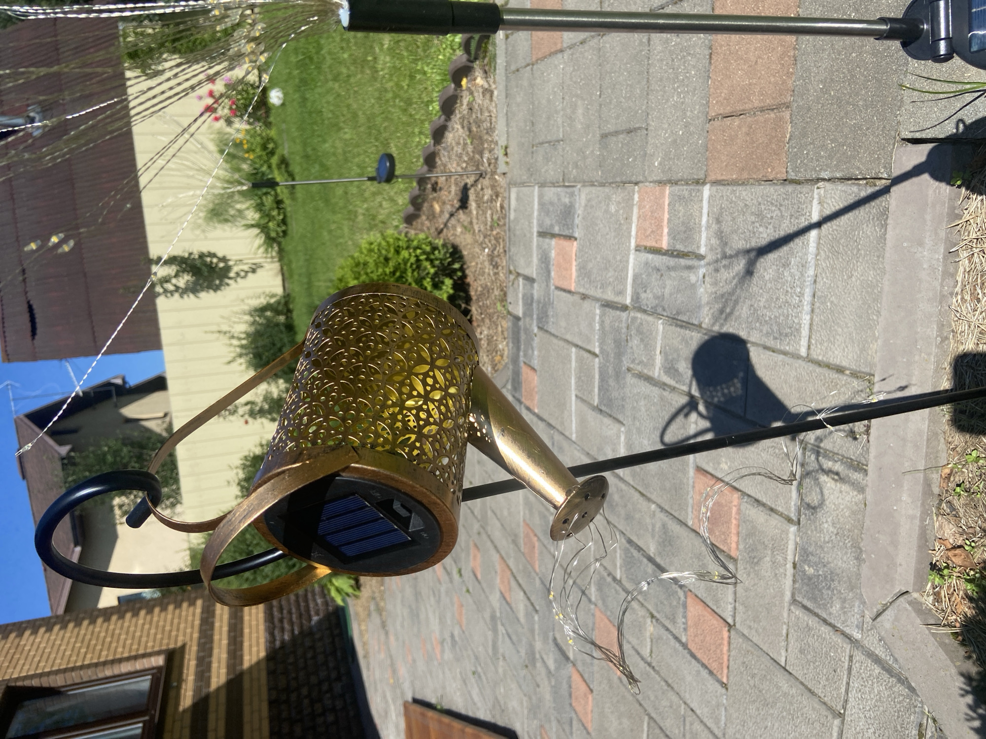 A decorative solar-powered watering can light fixture is displayed on a paved patio. The watering can is made of gold-colored metal with intricate cut-out patterns and a solar panel on its side. It is suspended by a black metal stand. Attached to the spout of the watering can is a string of small LED lights that trail downwards. The patio is made of gray and reddish-brown interlocking pavers. In the background, a grassy lawn is visible, bordered by a light-colored fence. A building with a brick facade and a brown roof is situated to the left. The scene is illuminated by bright sunlight, casting shadows on the ground. The location is identified as Зимородок Днп, Russia.