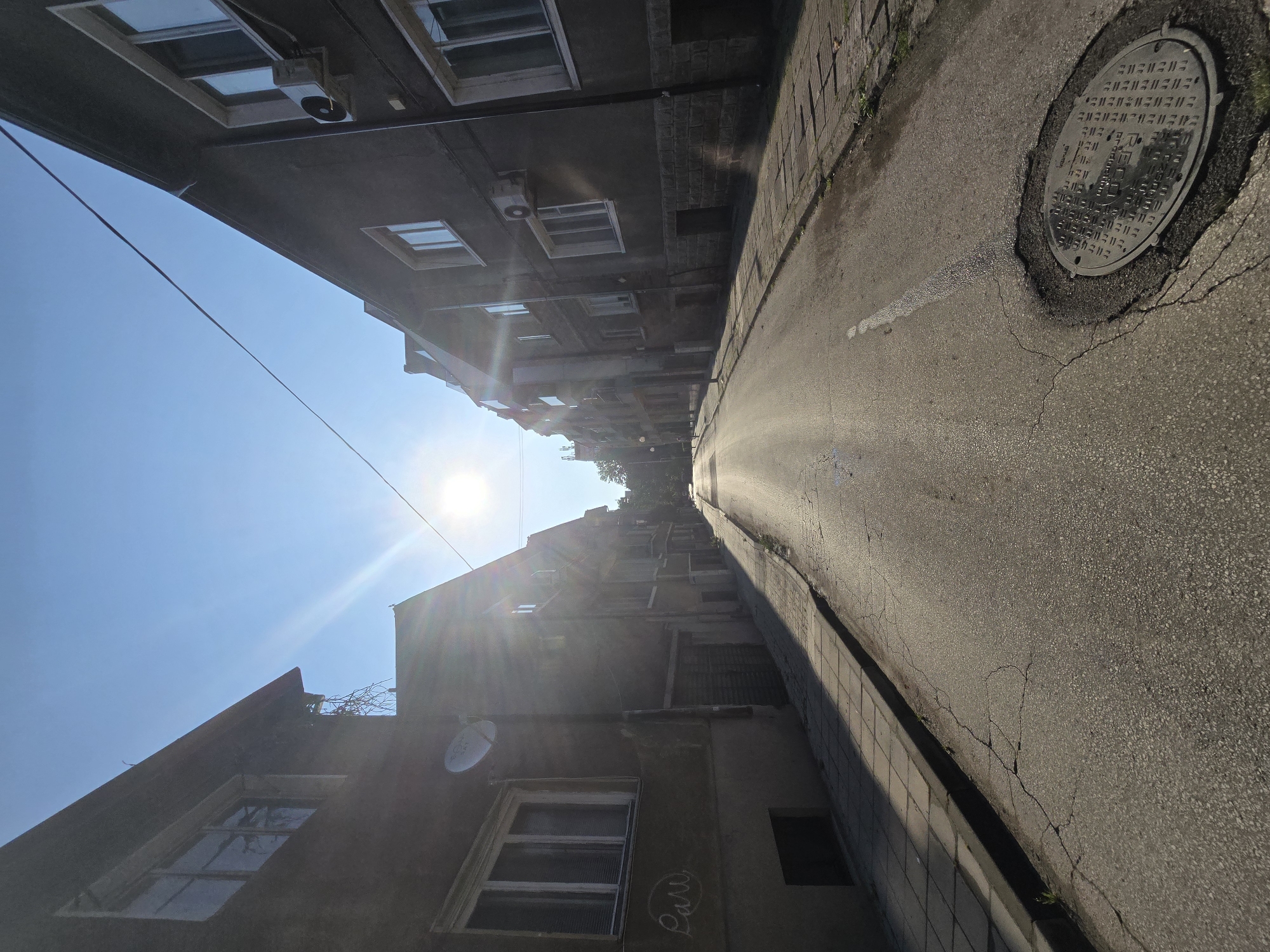 The image captures a narrow, sunlit street flanked by multi-story buildings under a clear blue sky. The perspective is tilted, making the street appear to curve sharply upwards along with the buildings on either side. The sun is bright and high, casting strong light and lens flare into the frame and creating distinct shadows along the street and buildings.
On the left side, several older buildings with multiple windows are visible. Some have external air conditioning units and one features a small satellite dish. Electrical wires stretch across the sky between the buildings. The building facades appear worn, with some visible weathering and patches. Faint, illegible graffiti can be seen near a window on one of the lower buildings.
The street itself is asphalt, showing signs of cracks and repair. A white line is painted along its length. A paved sidewalk runs alongside the street. A prominent feature on the street is a metal manhole cover, around which the asphalt is cracked. The manhole cover has Cyrillic text etched into its surface, clearly reading "ВК-СОФИЯ" (VK-SOFIA), followed by a grid-like pattern of characters.
The scene depicts a quiet daytime setting, likely in a city, with no human activity visible. The overall impression is one of a bright, ordinary urban environment with older architecture.
