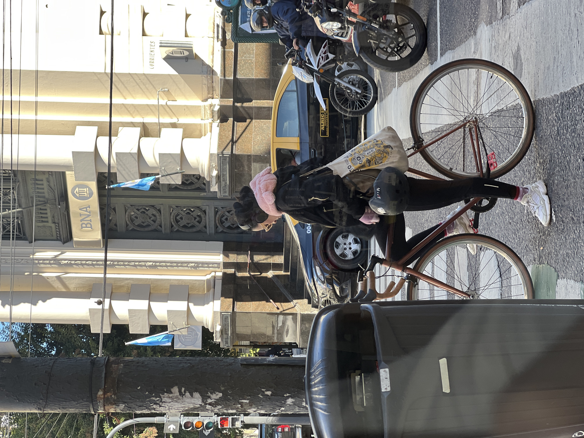 The image captures a street scene in Buenos Aires, Argentina. A person on a bicycle, wearing a pink head wrap, black leggings and white sneakers, is positioned on a crosswalk. A backpack rests on the bicycle's rear rack. The street is lined with parked motorcycles and a taxi. The building behind the cyclist is the Banco de la Nación Argentina (BNA) headquarters, located on Avenida Corrientes. A black post with a traffic light is visible on the left.