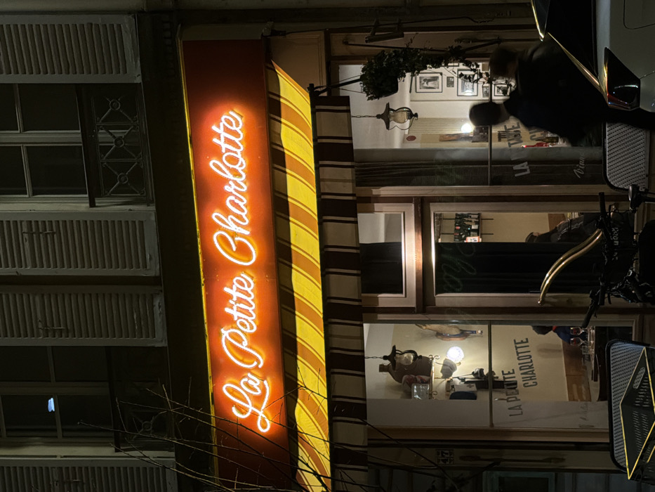 "La Petite Charlotte" glows in neon against an orange sign with yellow stripes. The restaurant's windows reveal interior scenes: framed pictures, a vintage lamp, and wine bottles. A person is partially visible in the top window, while a motorcycle reflects light in the foreground. A building with closed shutter windows sits next to the restaurant. Bare tree branches add a touch of winter to the night scene.