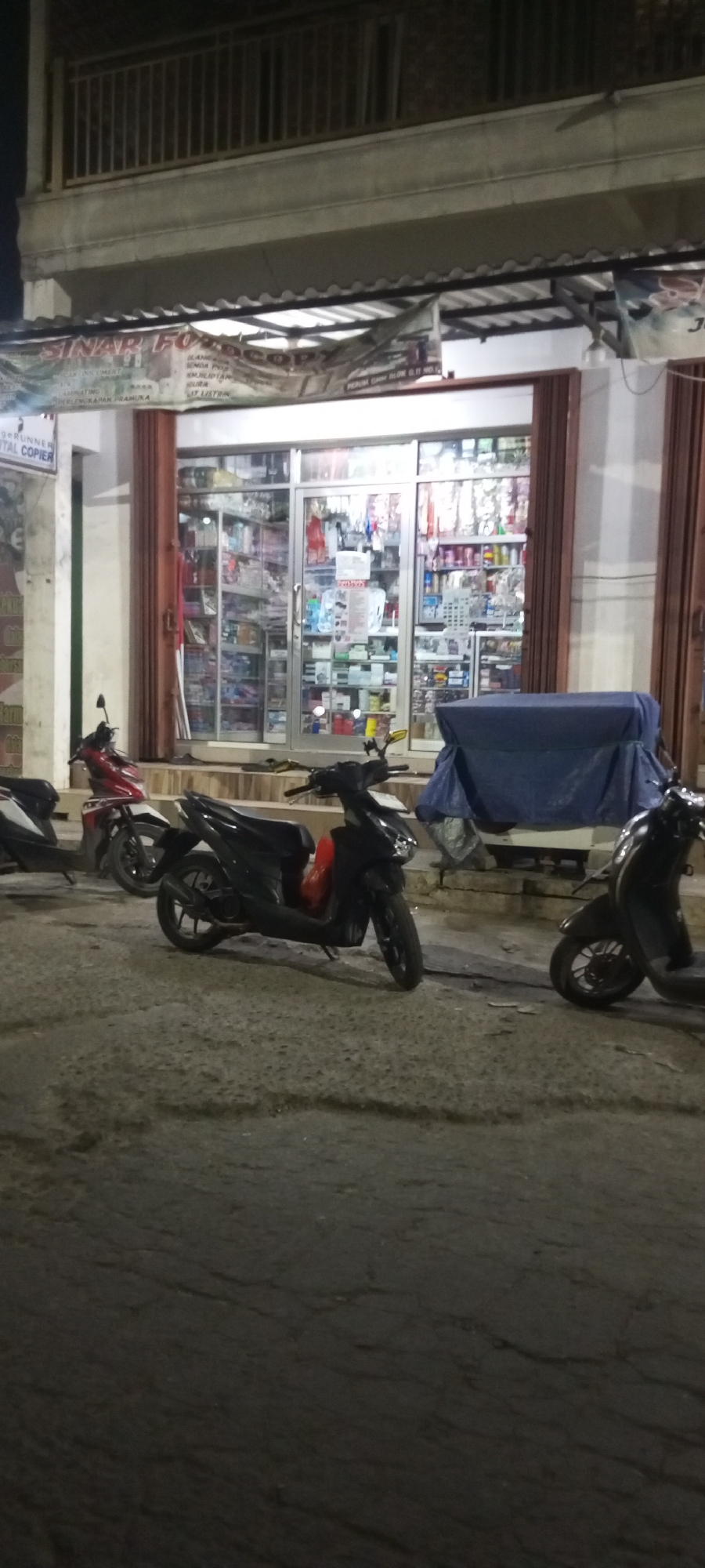 A wide shot captures a street-level view of a small business in Setu, Indonesia, at night. The business, identified by a banner as "SINAR FOTOCOPY," is a retail establishment with glass doors and windows displaying a variety of products. The interior is illuminated, revealing shelves stocked with goods. In front of the store, three motorcycles are parked on the paved street. One motorcycle, red and white, is positioned on the left. Another, black with a red accent on the side, is in the center, and a third, dark-colored, is on the right. A blue tarp covers an object behind the central motorcycle. The building appears to be a single-story structure with a balcony railing visible on the upper level. The lighting suggests it is nighttime, with artificial light illuminating the storefront and the immediate surroundings.