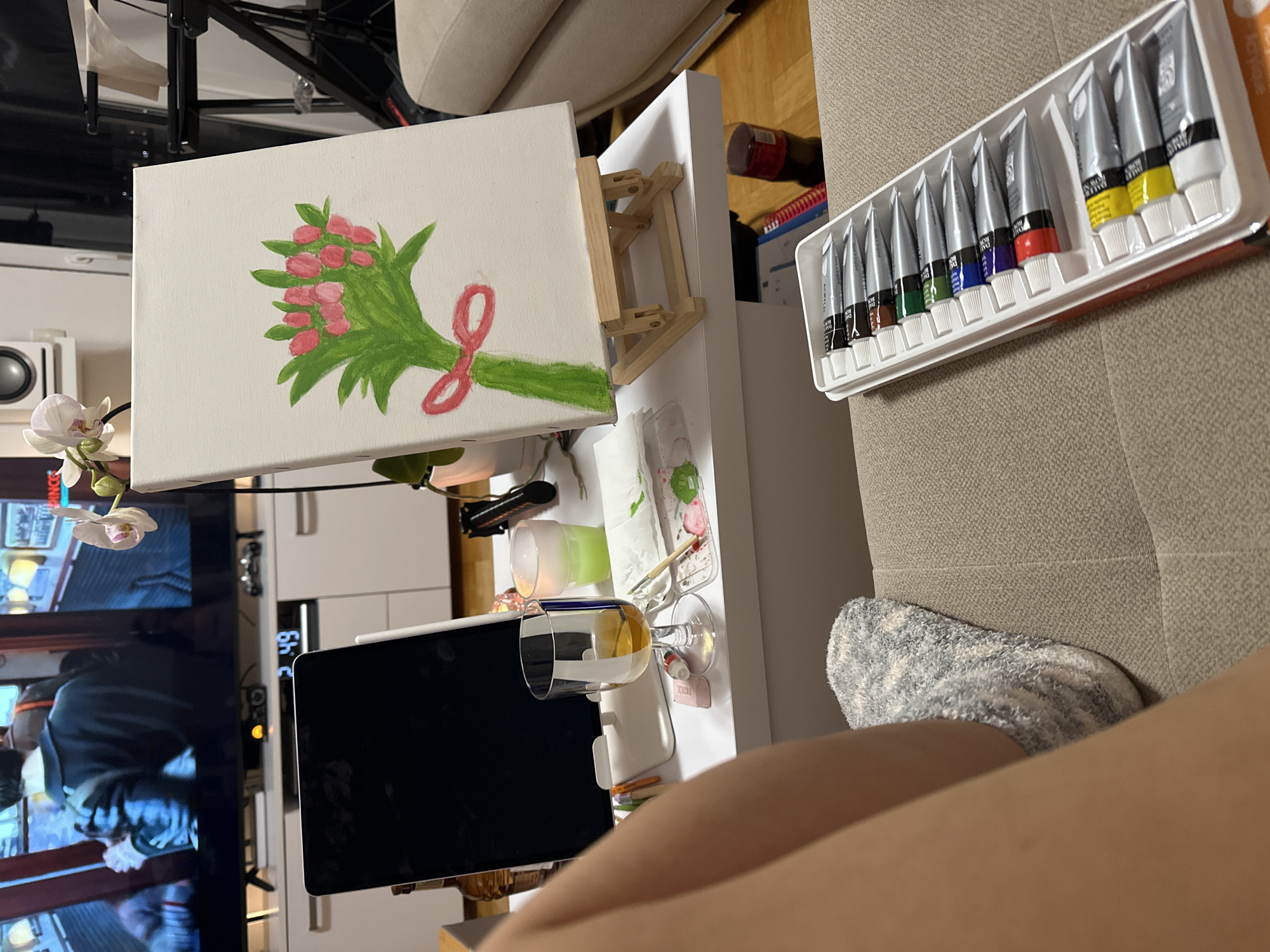 This image depicts an indoor living room scene in Budva, Montenegro, focused on a person engaged in a creative activity. In the foreground, the lower left leg and foot of an individual, wearing a grey and white patterned fuzzy sock, rest on a beige upholstered sofa. Adjacent to the person on the sofa, a white plastic tray contains a set of twelve acrylic paint tubes with various colored caps.
A white rectangular coffee table occupies the midground. On its left side, a dark-screened tablet stands vertically on a white holder, with a brown glass bottle partially visible behind it. Centered on the table, a clear glass holds an amber liquid, positioned near a small white rectangular object, a small pink paint tube, and a clear plastic palette with dabs of green and pink paint and two paintbrushes. To the right on the table, a small wooden easel supports a white canvas displaying a painting of a bouquet with pink, oval-shaped flowers, green leaves, and a red bow. Beside the easel, a small white ceramic pot holds a white orchid. Further right, a translucent green glass illuminates with a green glow, and a black cylindrical object rests nearby.
In the background, a large flat-screen television displays a movie or show scene featuring a person in a dark jacket seen from the back. Below the television, a white media console with closed cabinets shows a digital clock displaying "49". A white speaker is mounted on the wall to the left of the television. A light-colored sofa and a black tripod stand are partially visible in the far right background.