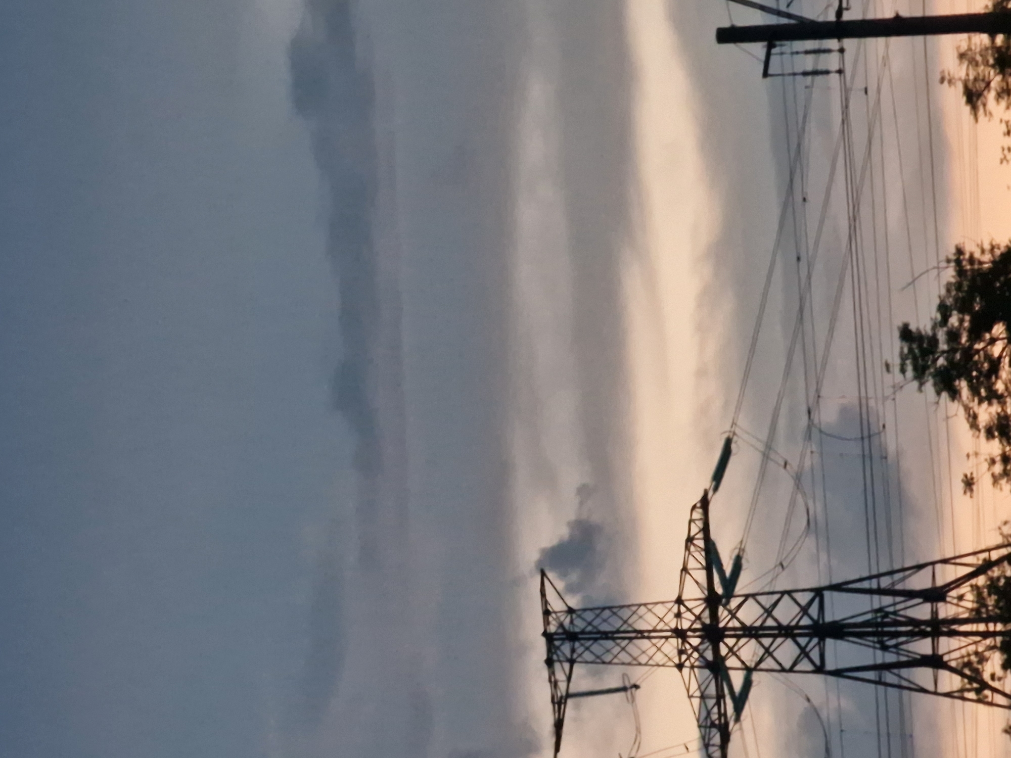 A tall, lattice steel electricity pylon dominates the left side of the frame against a cloudy sky. Multiple power lines stretch across the image from the pylon to smaller poles on the right. The sky transitions from dark grey clouds at the top to lighter, yellowish hues towards the bottom horizon. Silhouetted trees are visible at the bottom of the image, partially obscuring the ground. The scene appears to be set during dusk or dawn.