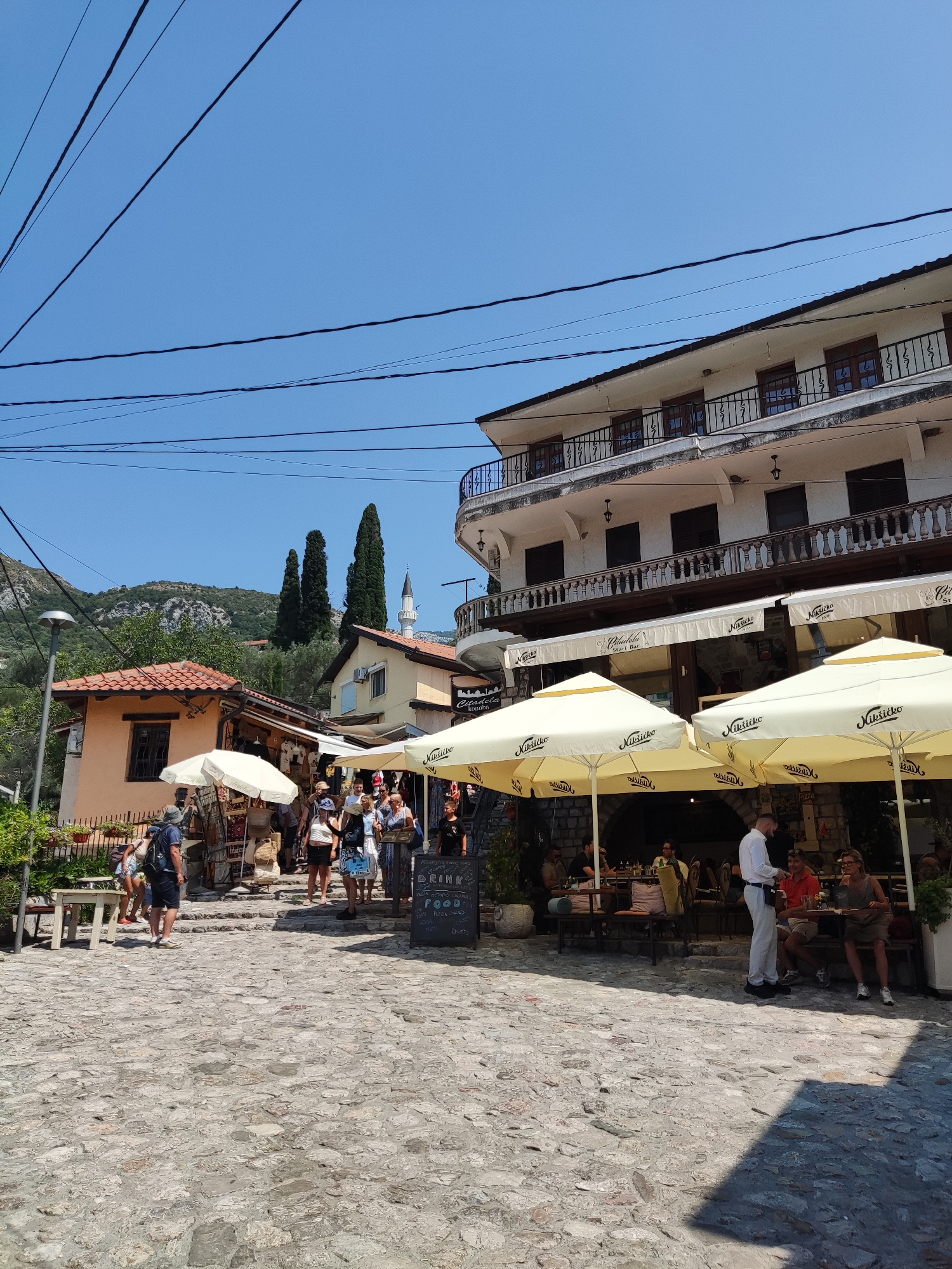 The image depicts a bustling outdoor scene in the city of Stari Bar, Montenegro. The foreground features a cobblestone street lined with shops and restaurants. Several people are walking along the street, browsing goods displayed outside a shop, and sitting at outdoor tables of a restaurant. The restaurant has large yellow umbrellas providing shade for its patrons, and menus are visible on a blackboard. In the background, there are buildings with tiled roofs and balconies, as well as cypress trees and a mosque minaret. The sky is clear and blue, suggesting a sunny day. The overall impression is one of a lively and historic urban environment.