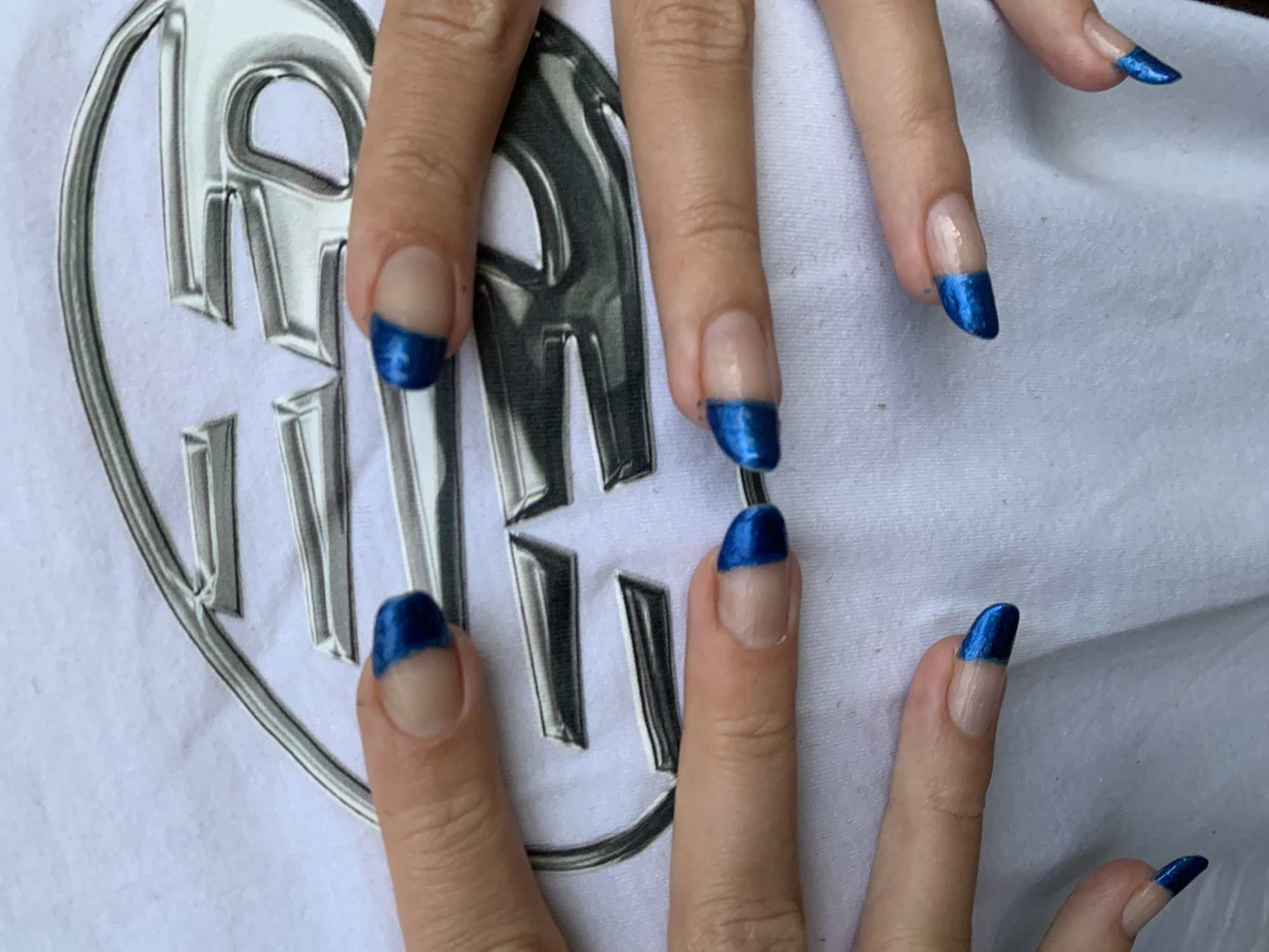 The image shows a close-up view of a person's hands with painted blue French tip fingernails. The hands are positioned over a white t-shirt with a silver metallic logo design. The scene does not depict anything indicating the city of Barcelona, Spain.
