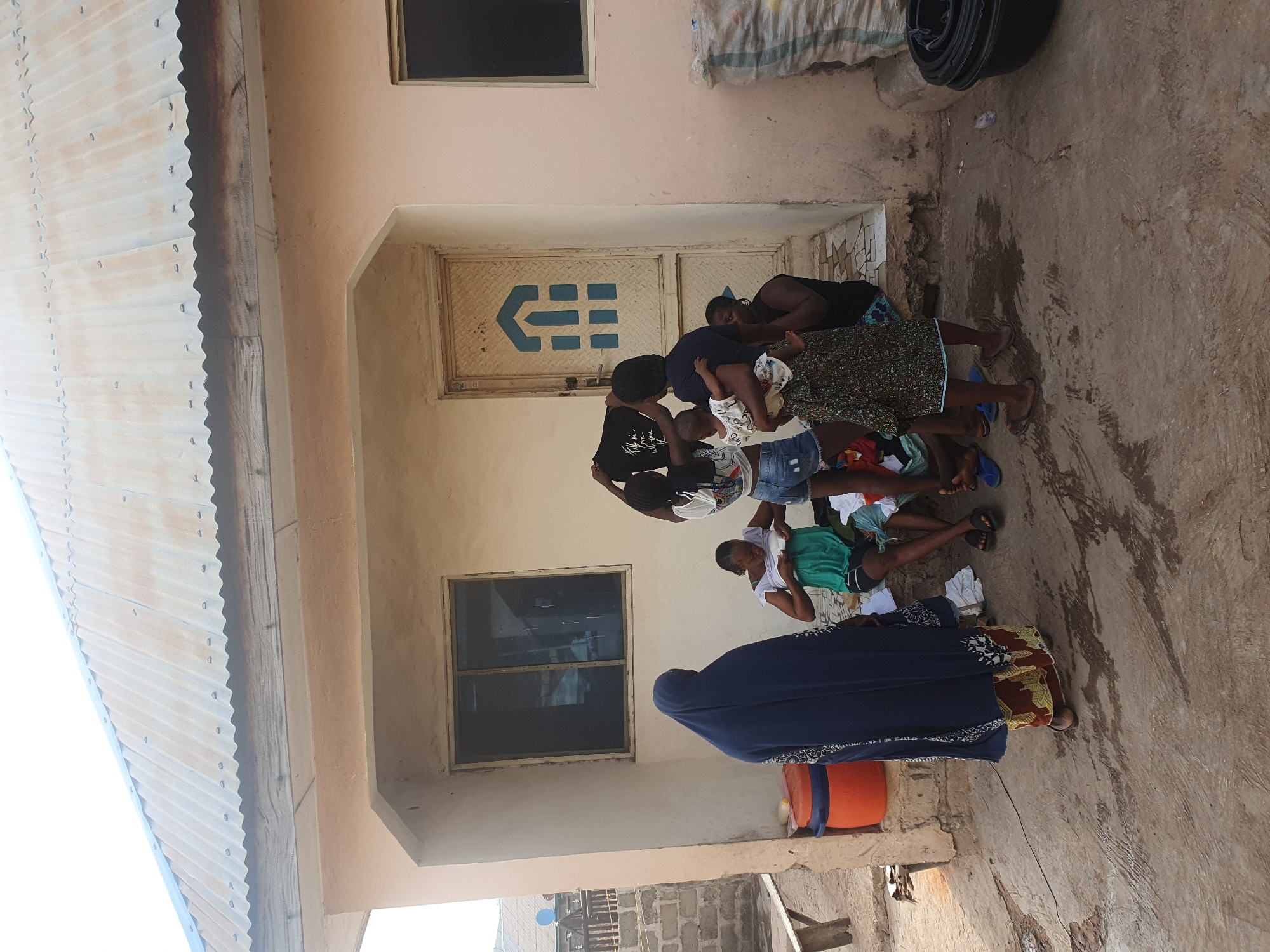 The image depicts a gathering of people, primarily women and children, in what appears to be a courtyard or veranda of a building in Lokoja, Nigeria. The scene is illuminated by bright daylight, suggesting midday or early afternoon.
In the foreground, a woman wearing a long, dark blue abaya-style garment with white embroidered patterns and a head covering stands with her back to the camera. Near her is an orange bucket. Further back, a group of women and children are clustered near a building with light-colored walls and a corrugated metal roof visible on the left.
Central to the group is a large metal door featuring a prominent blue, stylized arrow-like symbol or logo, potentially resembling "CHE," pointing to the left with horizontal lines inside. Several individuals are seated or standing around this area. One woman in a dark patterned skirt is seen with children, and another woman in a white t-shirt and dark shorts is holding a baby. A person in a black t-shirt is also present among the group.
The ground is a worn, dirty concrete surface. In the upper right corner of the image, stacked black plastic basins and a large fabric sack are visible. Windows with dark panes are set into the building's walls. The overall impression is a casual, communal scene in an outdoor domestic setting.