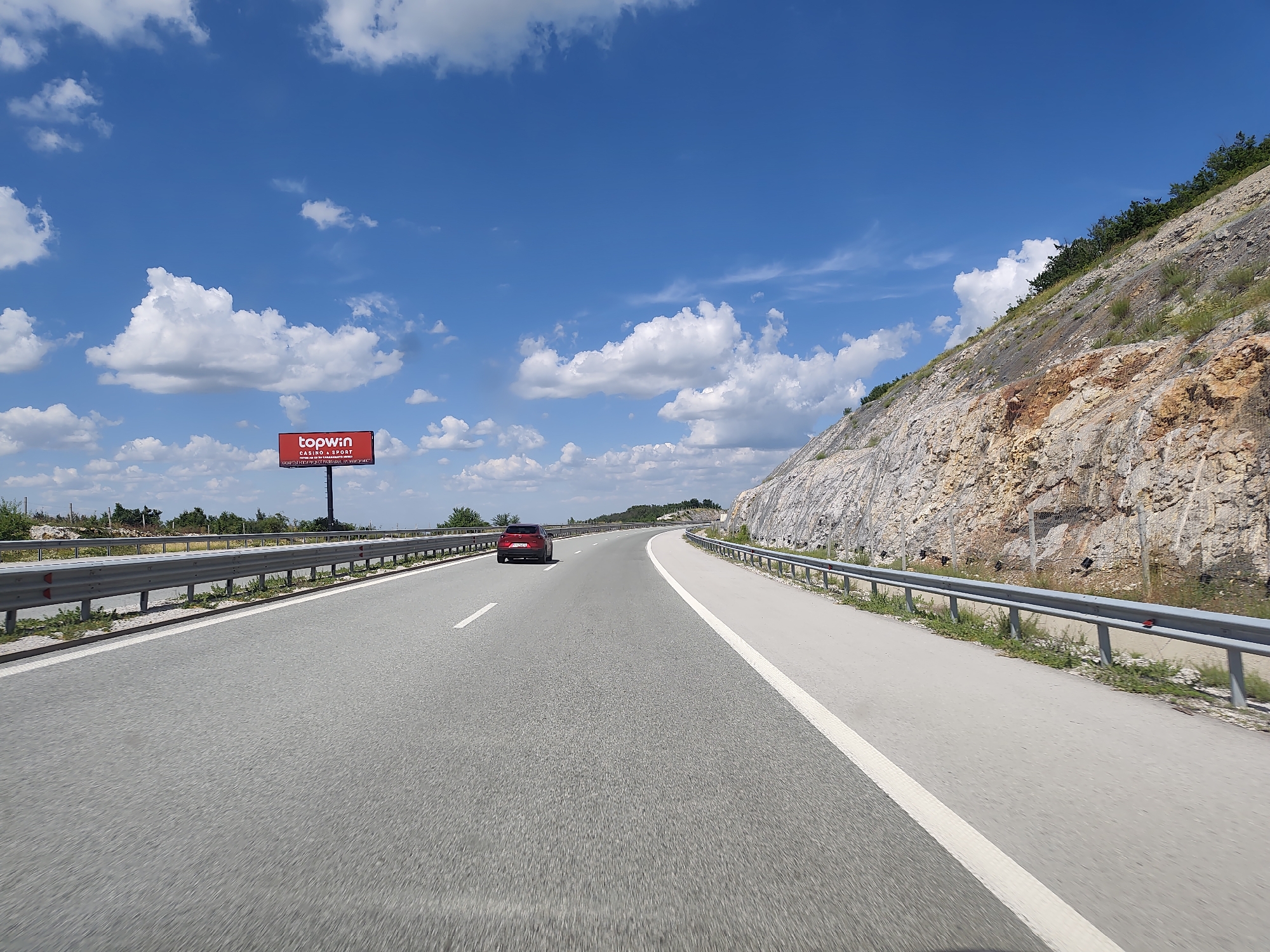 The image depicts a view from a vehicle traveling on a highway, likely near Yablanitsa, Bulgaria, given the context. The highway is paved with asphalt and has two lanes in the direction of travel, separated by a white line. A red car is visible in the distance, traveling in the same direction. To the right of the highway, a steep, rocky hill rises, with sparse vegetation and some trees at the crest. A metal guardrail runs along the edge of the highway, separating it from the embankment. On the left side of the highway, a large billboard advertising "topwin CASINO & SPORT" is visible, featuring a red background and white text. The sky is blue with scattered white cumulus clouds. The overall scene suggests a sunny day on an open road with natural and man-made elements in the landscape.