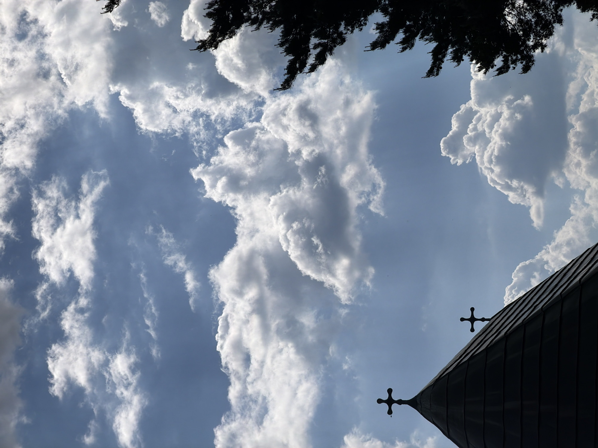 The image presents a low-angle view of the sky with cumulus clouds and the corner of a dark building with two crosses mounted on its roof. Some tree foliage is visible at the top. The location may be Otilovići, Montenegro, based on the request.