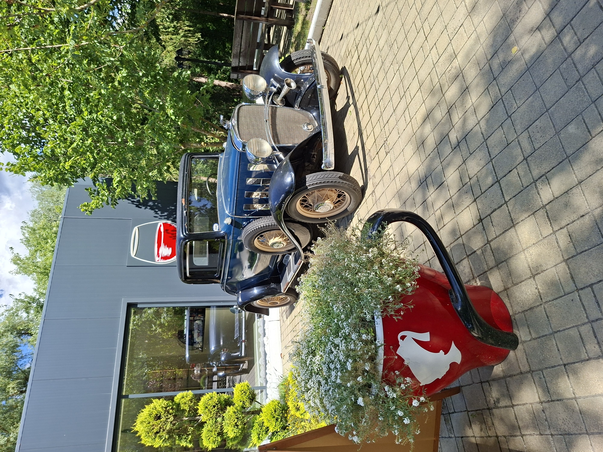 In Milestii Mici, Moldova, a dark blue vintage coupe with a chrome grille and spoked wheels is parked on a paved area. The car has a spare tire mounted on the side. In the foreground, a large red planter shaped like a cup, decorated with a white silhouette of a head wearing a crown, overflows with small white flowers. Behind the planter and to the left of the car is a modern building with large windows reflecting green trees. To the right of the car, more trees and a wooden bench are visible in the background. The scene is bathed in sunlight, casting shadows across the paving stones.