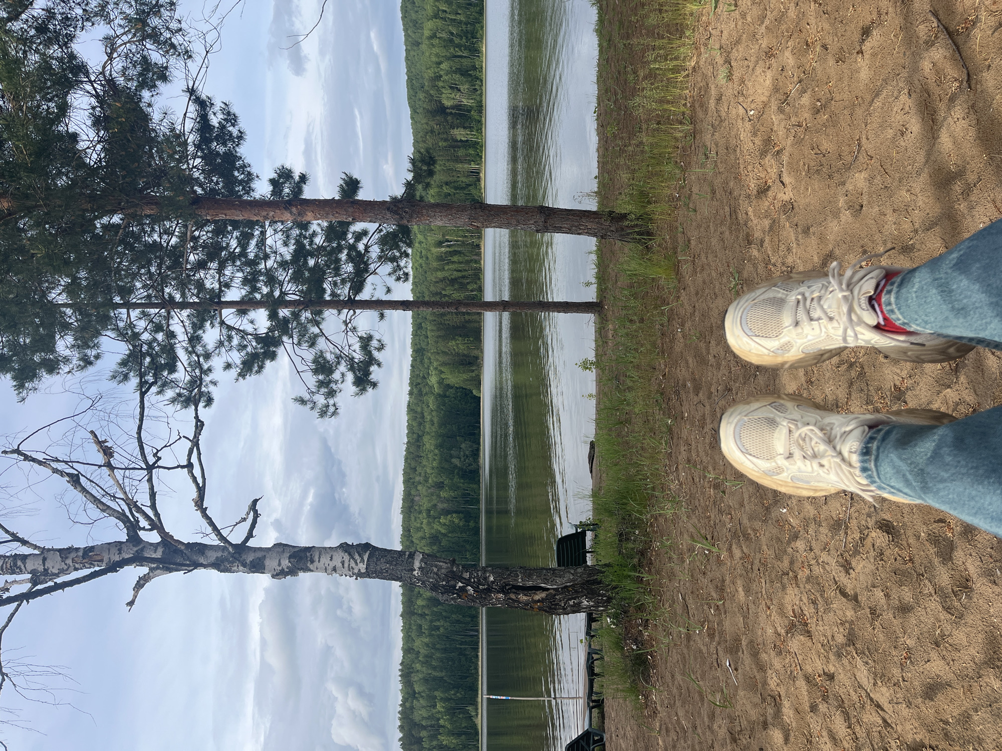 The image presents a landscape view captured at an outdoor location, specifically near a body of water, possibly a lake, judging by the presence of trees lining the shore and the expansive water surface. The foreground features a sandy ground area where two shoes are visible, indicating the presence of a person. The shoes are white trainers and appear to be worn, suggesting some activity. The person is wearing denim jeans. The backdrop consists of lush greenery and sky with some clouds. It is unclear from the image the exact location within the city of Unknown, Unknown. There are fishing chairs near the lake.