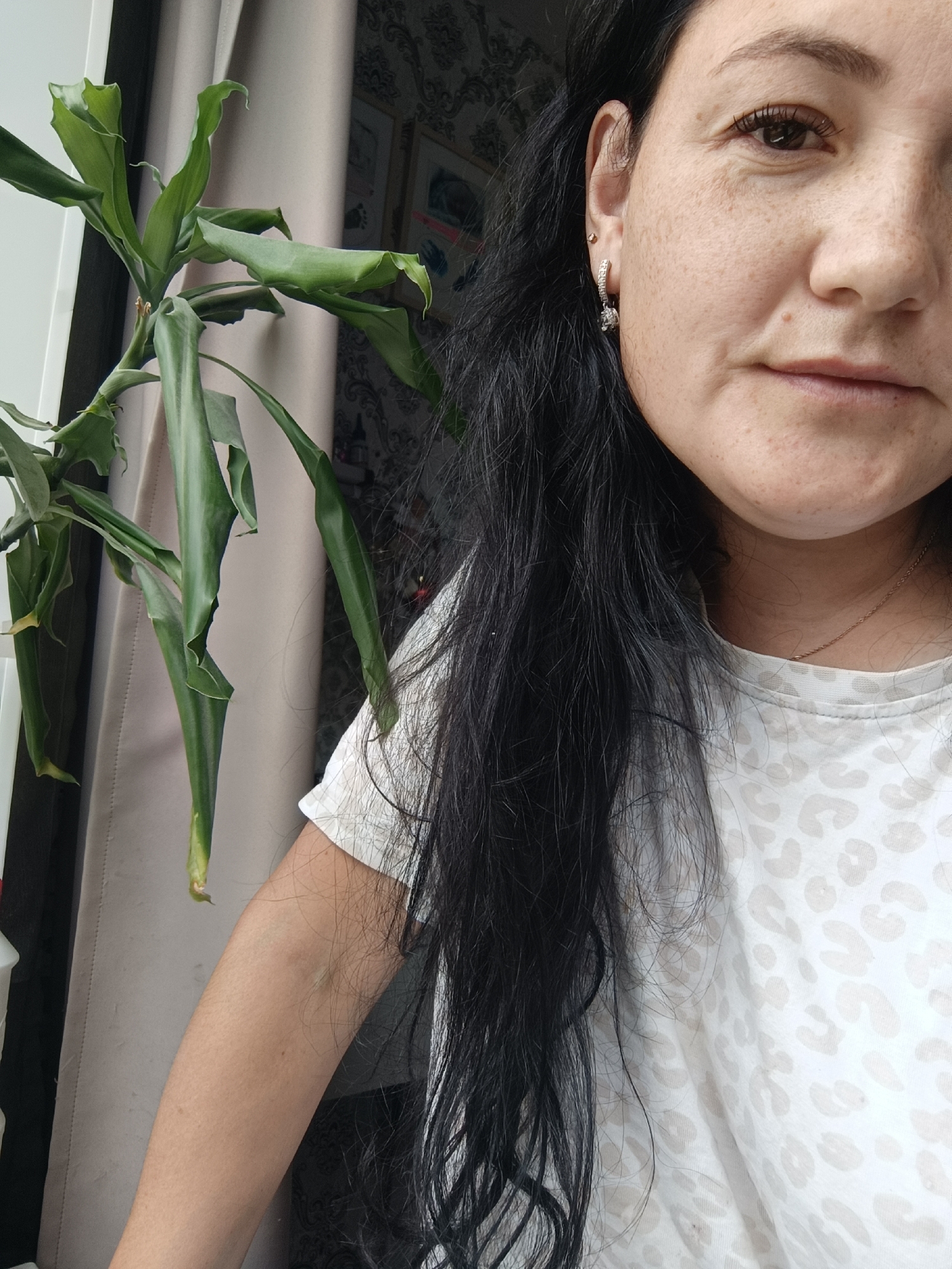 The image shows a close-up, waist-up shot of a woman with long, dark, wavy hair, wearing a white t-shirt with a light brown leopard print. She has fair skin with freckles and is wearing earrings in her left ear. A potted plant with long, green leaves is visible to her left. The background consists of a light-colored curtain and patterned wallpaper. The image appears to be taken indoors, possibly in a residential setting. There is no indication of the city of Chaikovsky, Russia, within the image itself.