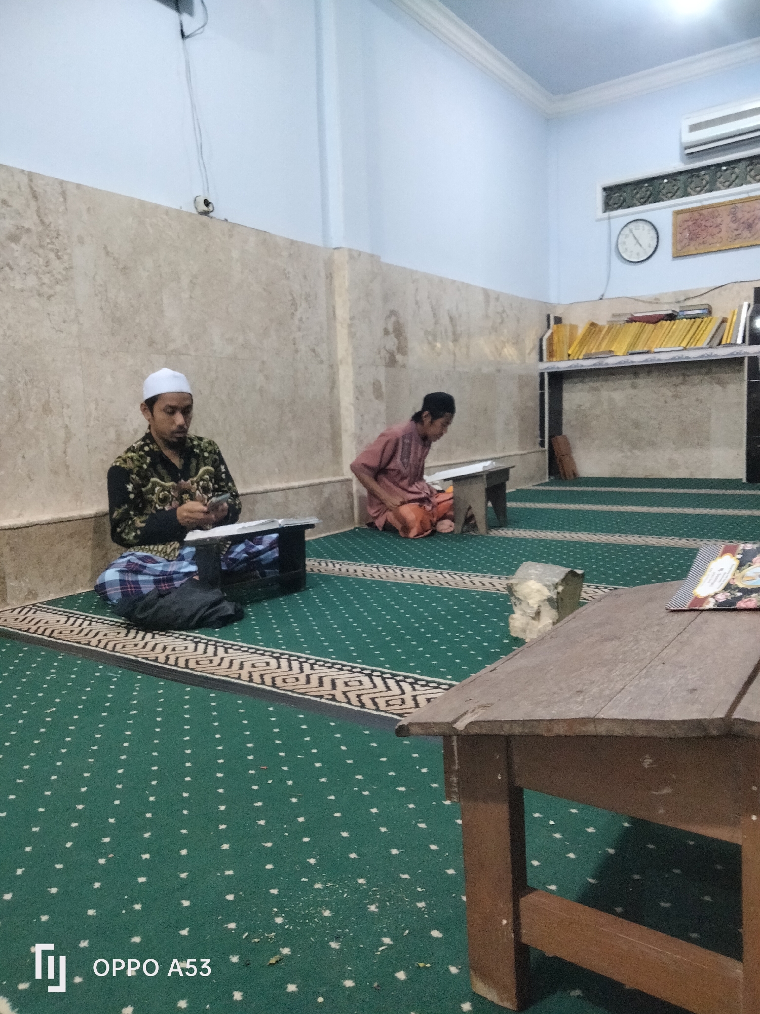 Two men are seated on a green carpet with a white and black patterned border in what appears to be a mosque or prayer room. The man on the left is wearing a white skullcap and a black and gold patterned shirt with a blue and white checkered sarong. He is looking down at a book on a dark wooden stand. The man on the right is wearing a peach-colored robe and a black skullcap. He is also looking down at a book on a similar wooden stand. In the background, there is a wall with a marble-like pattern, a shelf filled with yellow books, and a framed picture on the wall. A clock is also visible on the wall. A wooden table is in the foreground, partially obscuring the view of the carpet. The lighting suggests it is indoors, possibly at dusk or during prayer time. The overall scene conveys a sense of quiet devotion and study.