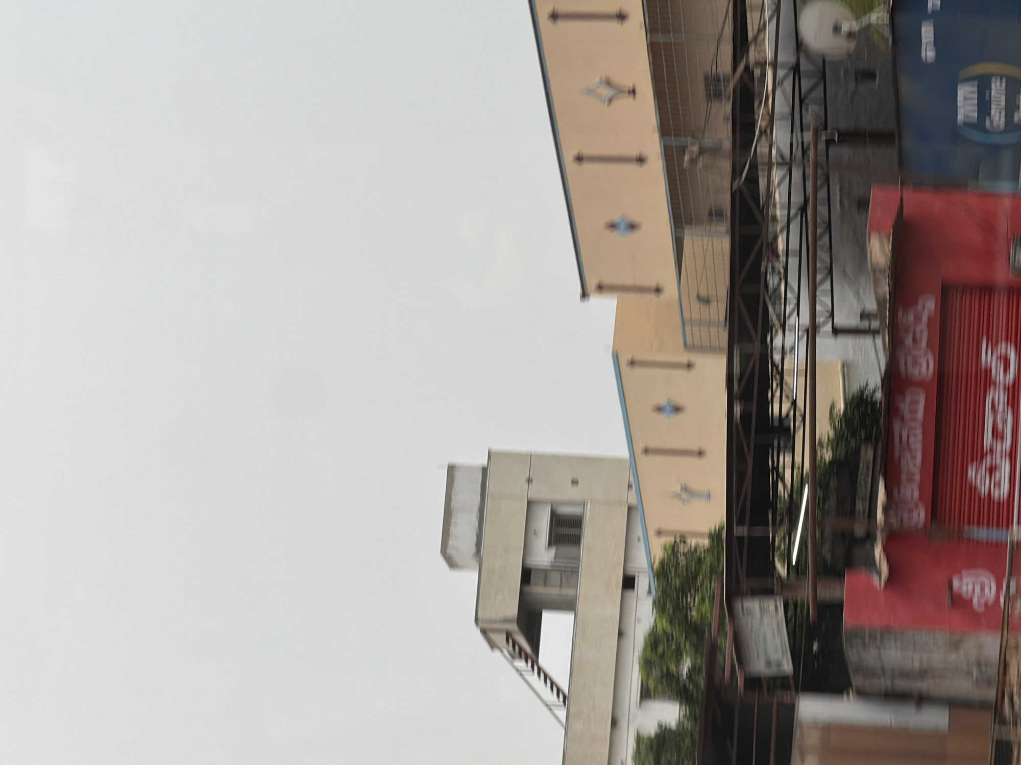 The image shows a street scene in Bethamcherla, India. In the background, there are several multi-story buildings. The building on the left is beige and has an external staircase leading to a rooftop area. It features several windows and a section with horizontal blinds. Adjacent to it is another building with a light brown facade adorned with decorative motifs that appear to be stylized diamonds and vertical lines. In the foreground, there are various structures, including a red building with a corrugated metal shutter and white text in a non-Latin script. To the right of the red building is a blue structure with text that includes "TATA Genuine". Trees are visible between the foreground and background buildings. The sky is overcast.