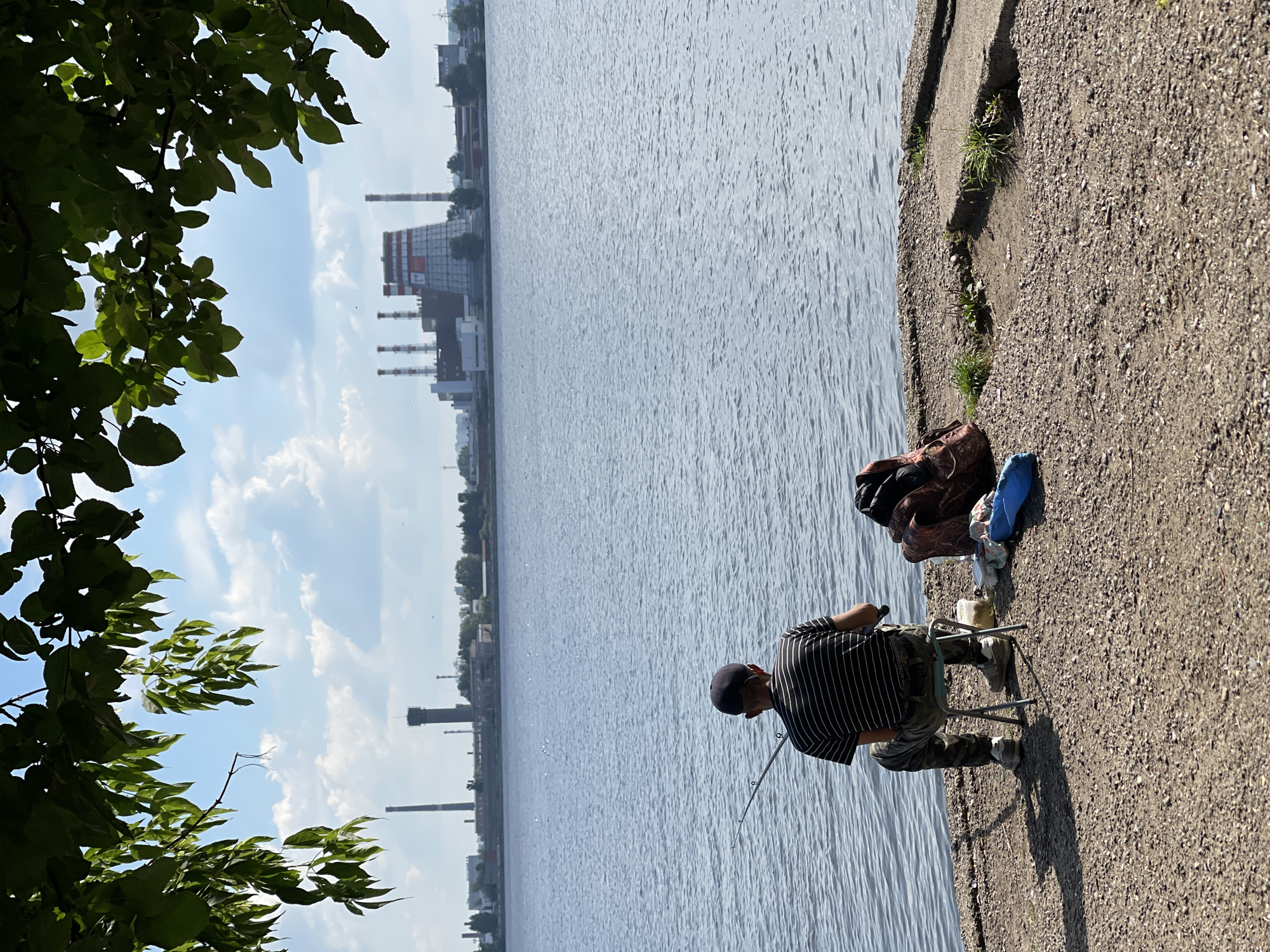 The image depicts a man fishing on the shore of a body of water, likely the Izh River, in Izhevsk, Russia. The man is seated on a small stool, facing away from the camera, with a fishing rod in his hands. Beside him, on the gravelly shore, are a backpack, a blue bag, and a small container. In the background, across the water, a distant industrial skyline is visible, featuring several tall smokestacks and large buildings, characteristic of a city's industrial district. The sky above is blue with scattered white clouds, and lush green foliage from overhanging trees frames the top of the image. The overall scene conveys a sense of calm and solitude, with the solitary figure engaged in recreational activity against a backdrop of industrial development.