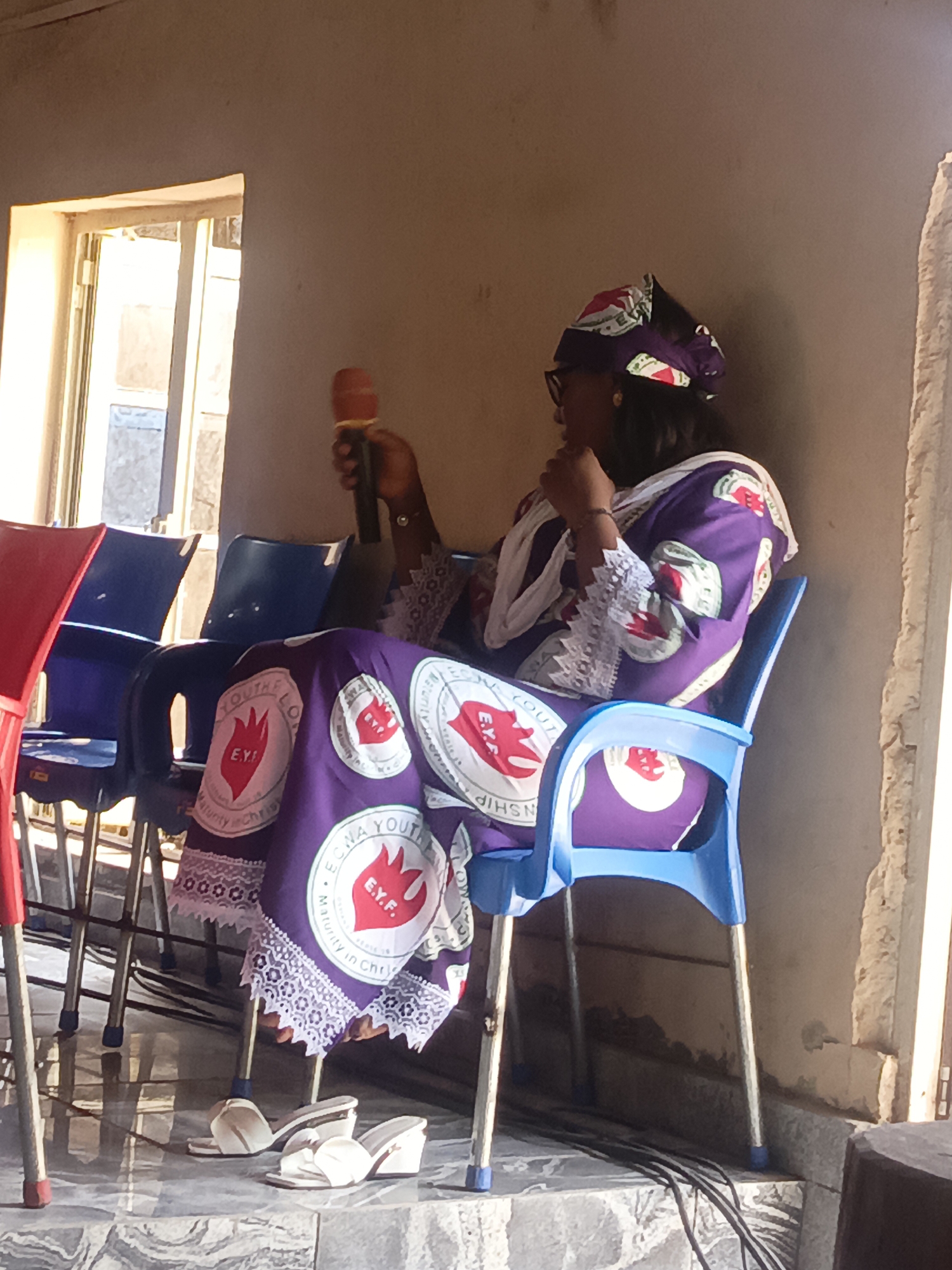 Akwanga, Nigeria: A woman sits on a blue plastic chair in what appears to be an interior space. She is holding a red microphone. The woman is dressed in a purple and white outfit with circular patches that read "ECWA YOUTH FELLOWSHIP" and the acronym "E.Y.F" around a heart-shaped logo. Her head is covered in matching material, and she is wearing glasses. She is also wearing white sandals. Behind her are several stacked blue and red plastic chairs. There is a window letting in daylight in the upper left of the image. Electrical wires are visible on the floor, near the base of her chair. The walls are painted a neutral color.
