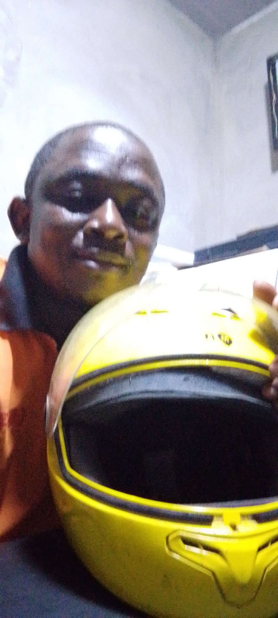 A man is holding a yellow helmet. The location is Lokoja, Nigeria. The setting appears to be indoors. The man is dark-skinned, and his head is partially balding. He is wearing an orange shirt. The helmet has a black visor opening. The background is a light-colored wall.