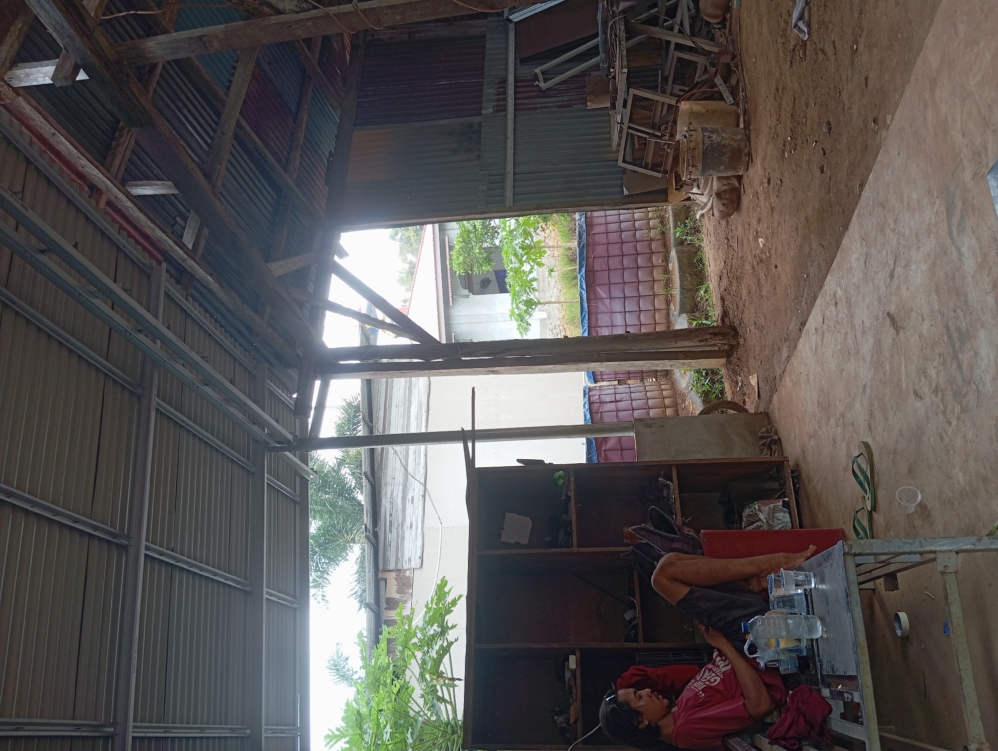 The image depicts an outdoor, covered workspace with a person sitting on a bench. The workspace appears to be located in Desa Retak Mudik, Indonesia, given the context. A man in a red t-shirt and dark shorts is seated on a metal bench, looking at a phone. In front of him on a metal table are several plastic cups, a bottle of water, and a glass of dark liquid. To his left is a tall, brown wooden shelving unit with various items stored on its shelves, including shoes and other miscellaneous objects. A red piece of cloth is draped over the back of the bench.
The environment is a rustic, open-air structure with a corrugated metal roof supported by wooden beams and metal framing. The floor is a mix of concrete and packed dirt. In the background, beyond the open structure, are glimpses of trees and buildings, suggesting a semi-rural or mixed-use area. To the right of the man, in the covered area, are stacks of metal frames and what appear to be industrial containers or drums, indicating a possible workshop or storage area. A large, dark water tank or pond with a checkered pattern is visible behind the shelving unit. The overall impression is a utilitarian and somewhat cluttered space.