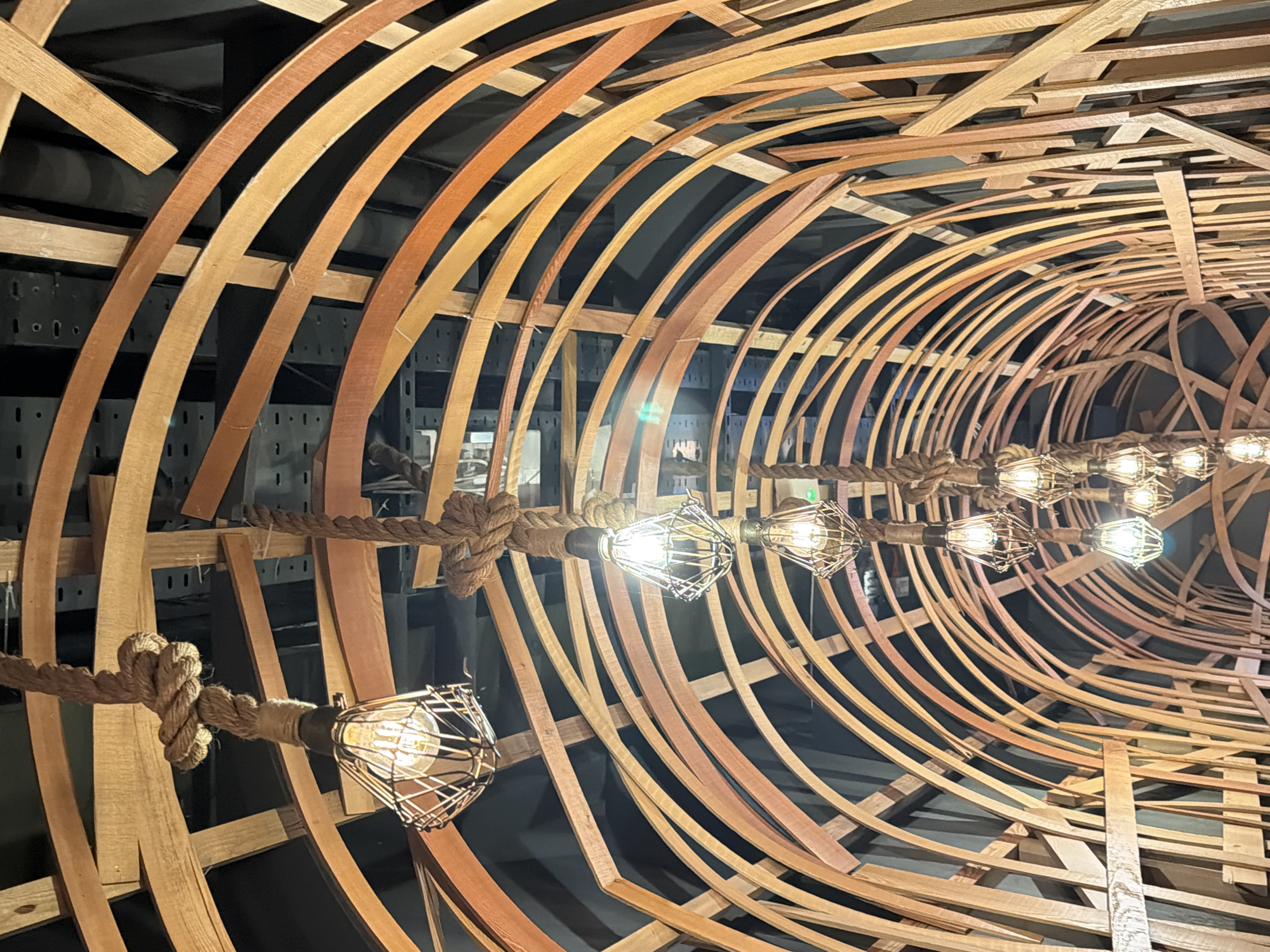 An intricate interior view showcases a distinctive tunnel-like structure constructed from numerous curved and straight wooden strips of varying warm brown tones, ranging from light tan to reddish-brown. These wooden elements are arranged to form a skeletal framework, reminiscent of the ribs of a boat hull or the staves of a large barrel, creating a sense of enclosure and depth.
Suspended through the center of this wooden armature is a thick, natural fiber rope, intricately knotted at intervals. From this rope hang multiple industrial-style light fixtures, each featuring a clear Edison-style bulb housed within a dark metal wire cage. These bulbs emit a warm, bright glow, casting light onto the surrounding wooden surfaces and enhancing their texture.
The background beyond the wooden structure is dark, revealing glimpses of dark metal shelving or structural supports in certain areas. This suggests an indoor setting, possibly a commercial establishment like a bar, restaurant, or retail space, where the artificial lighting contributes to a specific ambiance. The overall design evokes a rustic, nautical, or handcrafted aesthetic.
The scene is located in London, United Kingdom. There is no visible text in the image.