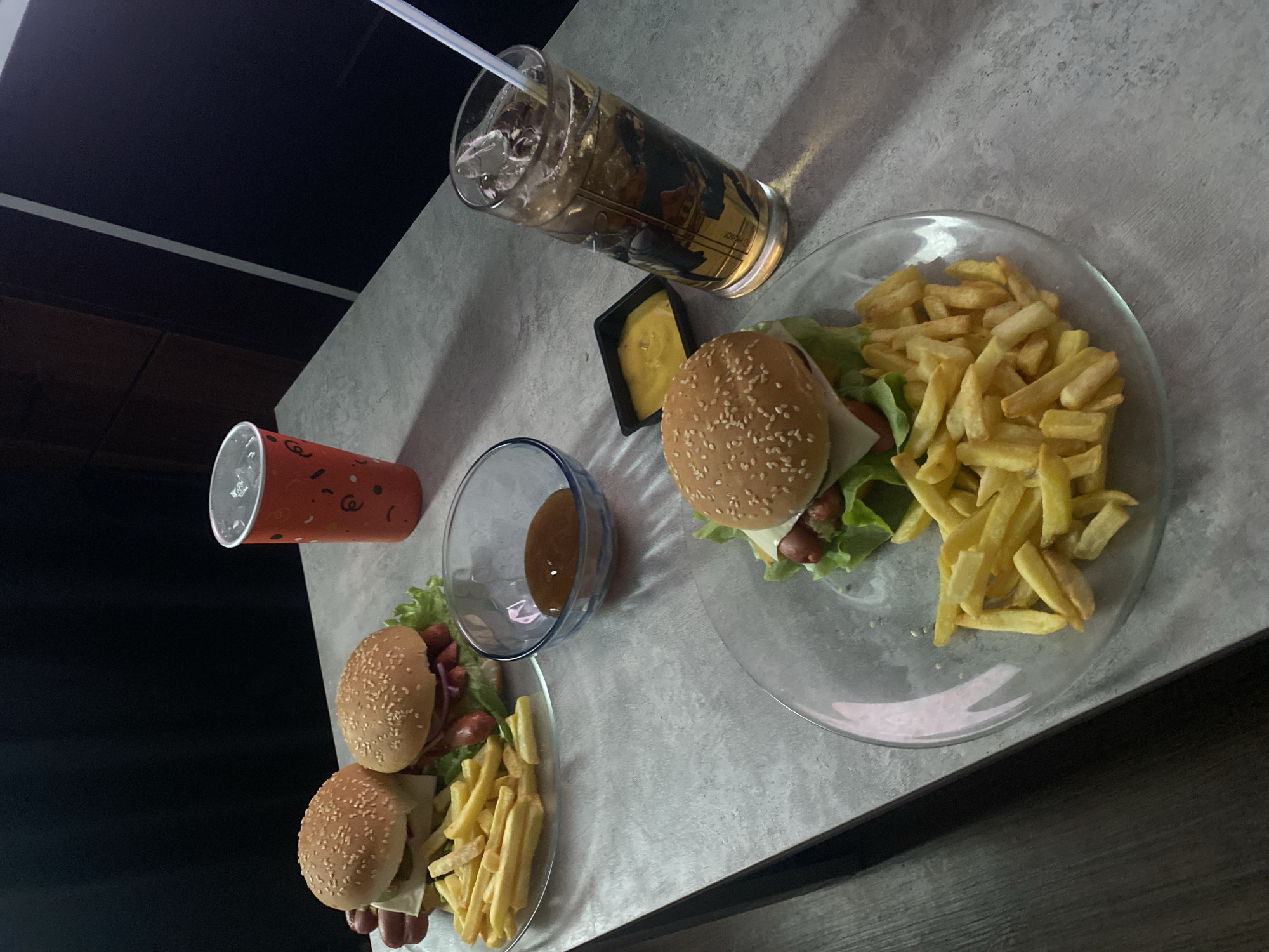 The image depicts a meal setting on a table, likely indoors, with fast food items. There are two plates, each containing burgers and french fries. A glass of iced drink with a straw and a small container of yellow sauce are present near one of the plates. A red paper cup and a small bowl of brown liquid are placed near the other plate. The overall presentation suggests a casual dining environment. There is no information to confirm that the location is Izhevsk, Russia.