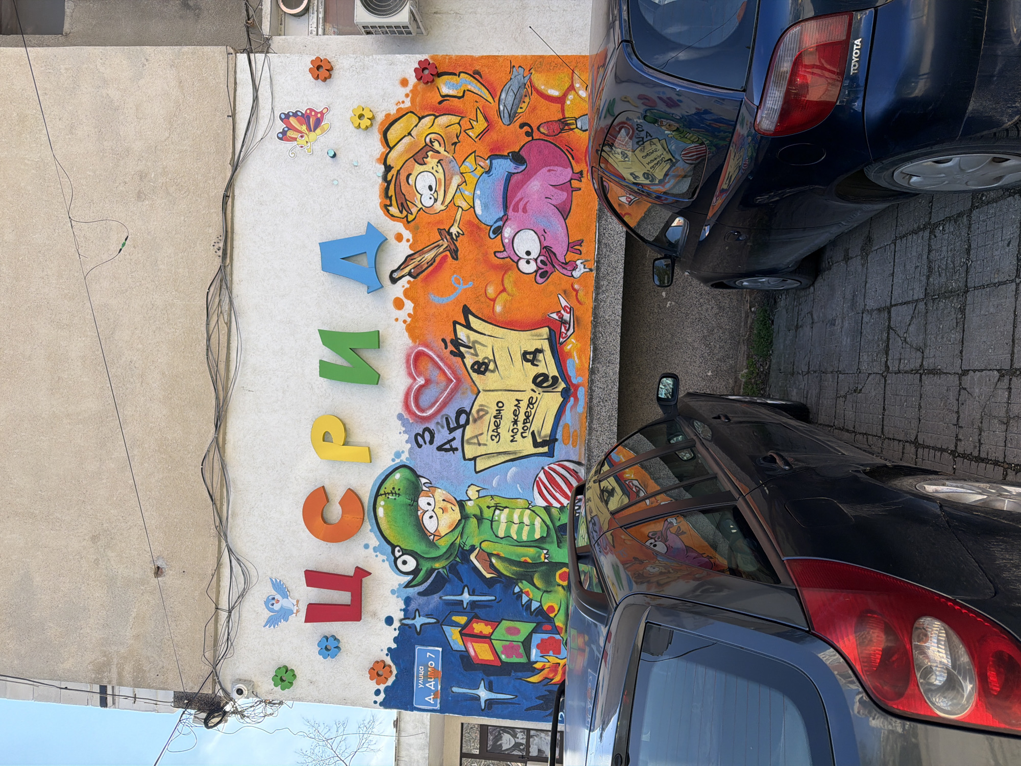 The image shows a mural painted on a building wall, partially obscured by two parked cars. The mural has colorful cartoon-style illustrations and Cyrillic text. The text includes the word "деца" (children) spelled out in large, separate letters and the phrase "Заедно можем повече!" (Together we can do more!). A sign reads "улица Д. Димо 7" (street D. Dimo 7), pinpointing the location in Pleven, Bulgaria. An air conditioning unit is attached to the wall above the mural. The cars parked adjacent to the mural are a blue Toyota and a dark gray car.