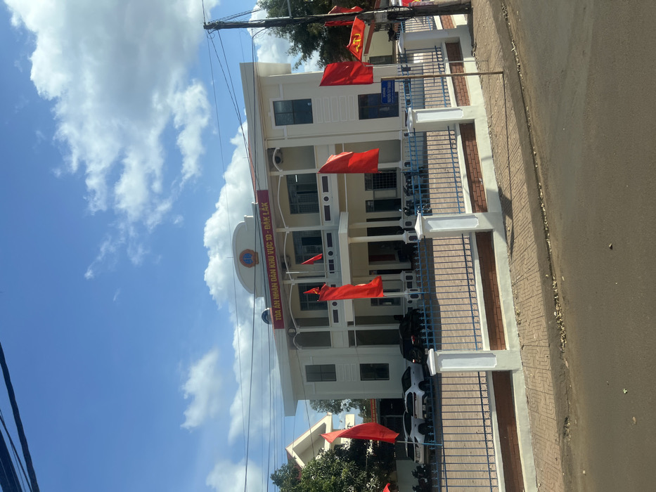 This image depicts the exterior of a two-story institutional building, identified by the banner "TÒA ÁN NHÂN DÂN KHU VỰC 10 - ĐẮK LẮK" (People's Court of Area 10 - Đắk Lắk), located in Trung Hồ, Vietnam. The building is light cream or off-white with a central entrance featuring columns supporting an upper balcony, above which a judicial emblem of scales is visible. Numerous rectangular windows are set into the facade.
In front of the building, multiple red flags on poles are displayed; one on the far right includes the yellow hammer and sickle emblem. Behind a blue metal fence with white concrete pillars and a brick-patterned base, several vehicles are parked: two white SUVs, one dark sedan, and multiple motorcycles/scooters.
The scene is under a clear blue sky with scattered white cumulus clouds. In the immediate foreground, a paved sidewalk with a geometric pattern separates an asphalt road from the fence. A utility pole with power lines is visible on the right side of the frame, alongside a partially visible blue street sign bearing Vietnamese text, likely "ĐƯỜNG NGUYỄN TẤT THÀNH." Green foliage and parts of another building are present in the background to the left. No individuals are clearly discernible.
