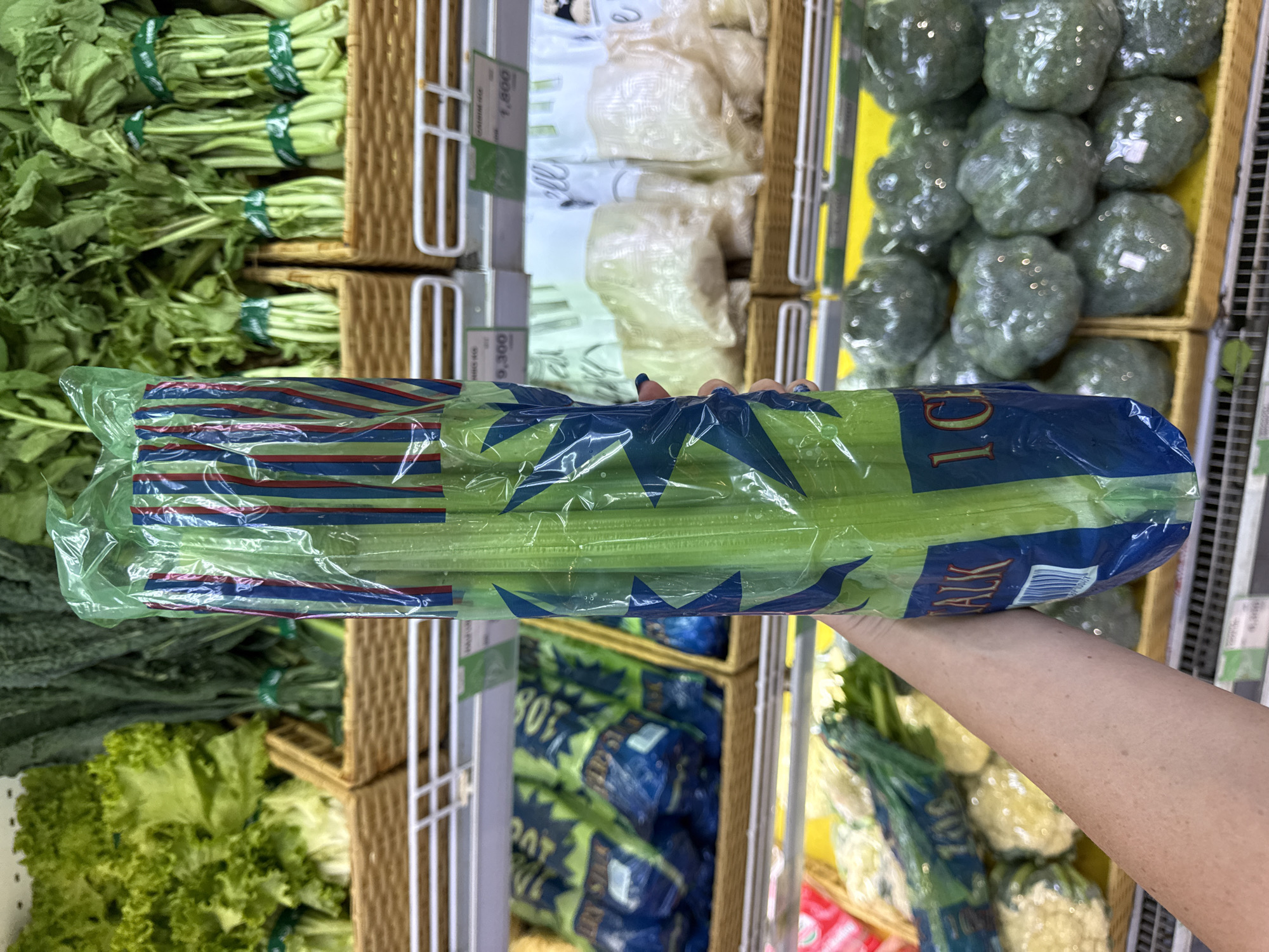 In Denpasar, Indonesia, at a supermarket, a person's arm and hand are prominently featured in the foreground, holding a large bunch of celery. The celery is enclosed in clear plastic packaging with vertical blue, red, and green stripes and blue star accents. Text on the packaging is partially visible, including "TALK" and "1. CE". Behind the celery, shelves display a variety of fresh vegetables. On the left, leafy green lettuce is visible in a woven basket. Further back, bunches of dark green leafy vegetables are also in woven baskets. In the middle ground and background, other produce items are on display, some in plastic packaging. The lighting suggests an indoor retail environment.