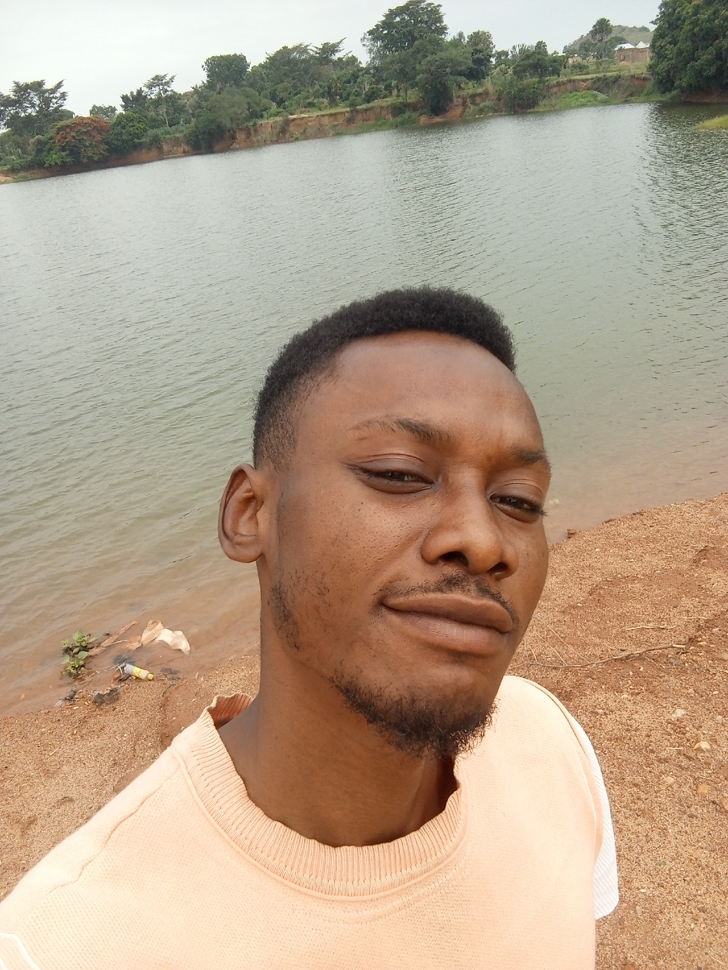 A close-up selfie of a young, dark-skinned man with short, dark hair and a goatee, standing outdoors. The background shows a body of water, possibly a lake or river, with trees and vegetation on the opposite bank. The foreground is a sandy or earthen shoreline. The man is wearing a light-colored sweater with white sleeves. There are signs of litter and discarded items at the water's edge. The location is Udu, Nigeria.