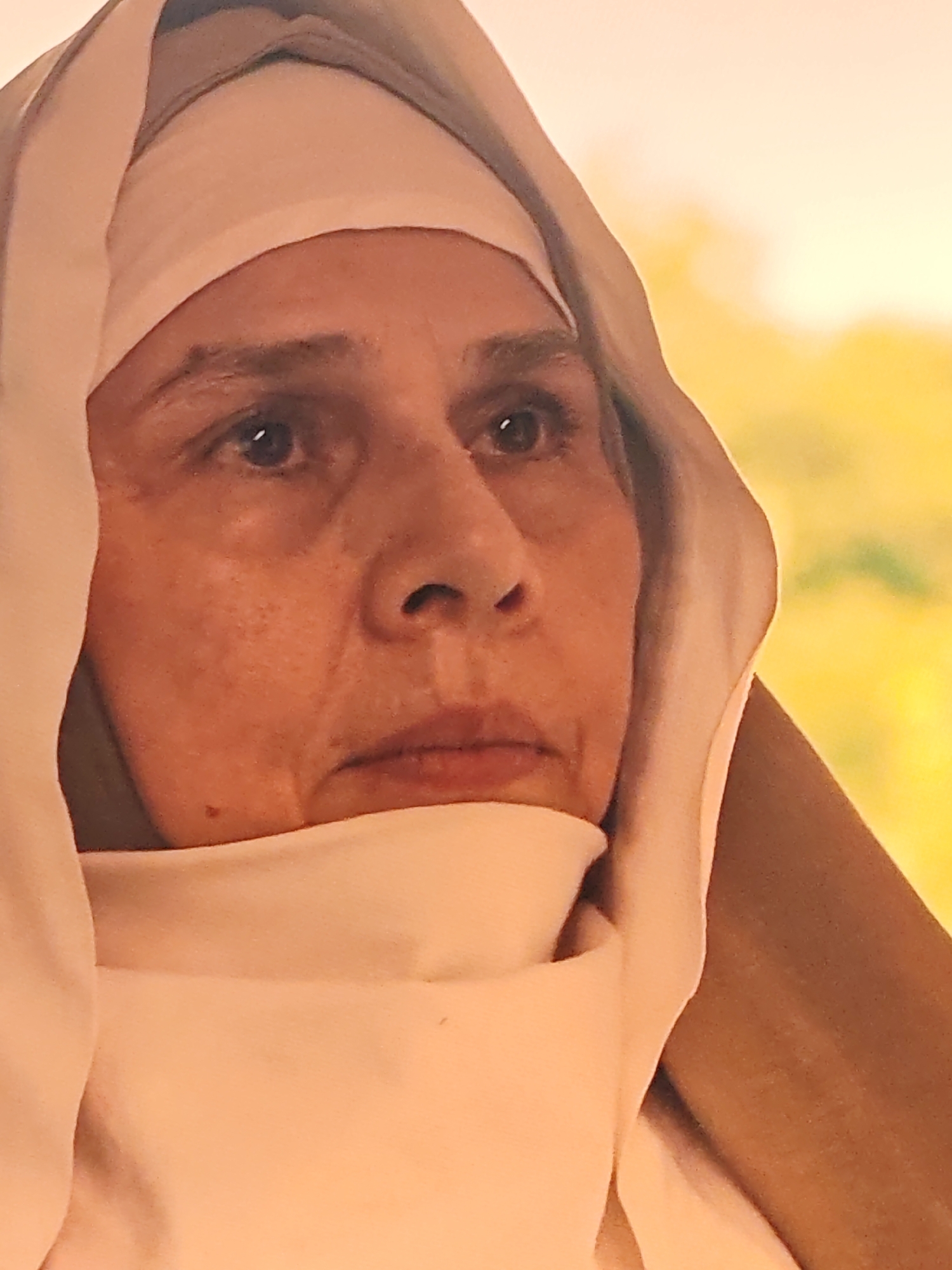 A medium shot captures the face of a woman wearing a white headscarf and habit, her head angled slightly to the right. The background is a soft, out-of-focus blur of yellow and green, suggesting a natural or dimly lit environment. The woman's features are somewhat aged, with lines around her eyes and mouth. Her expression is neutral, her gaze directed towards the viewer. The fabric of her habit is draped around her face and neck, framing her features. The lighting highlights the texture of her skin and the folds of her clothing.