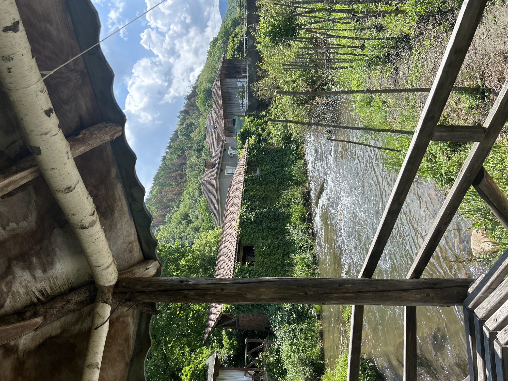This image captures a serene, rustic rural landscape on a bright, cloudy day, seemingly viewed from a sheltered vantage point.
In the immediate foreground, components of a wooden structure are visible, including thick, light-barked logs (resembling birch) serving as supports, dark wooden beams, and a corrugated roof section. This suggests the viewer is under a rustic shelter, possibly a porch or gazebo.
The middle ground features a clear, flowing river or stream with ripples visible on its surface. Along its right bank, a wire fence with wooden posts separates the river from cultivated or wild greenery. Beyond the river, several buildings are nestled amidst abundant foliage. One prominent building has a reddish-brown tiled roof, and its walls are partially covered in dense green ivy. Another structure further back appears to be an older, wooden building with a brown roof. An air conditioning unit is visible on the exterior of one of the buildings, hinting at modern amenities in a traditional setting.
The background is dominated by a lush, green hillside densely covered with trees and vegetation, rising under a bright blue sky with scattered white clouds. The scene conveys a sense of tranquility and natural beauty.
No people or animals are visible. The activity captured is a static view of this peaceful landscape. It is daytime, with clear evidence of sunlight. Some illegible text on signs is faintly visible on the wooden building in the mid-ground. The setting suggests a remote village or homestead within a mountainous or hilly region.