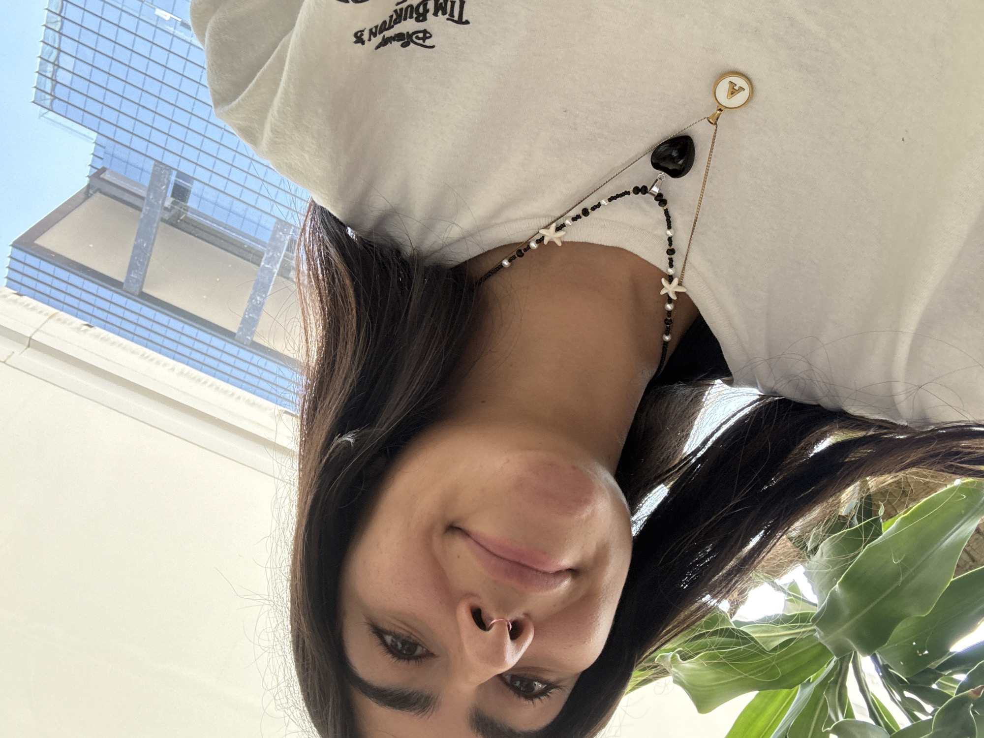 The image is a low-angle selfie of a woman with long, dark brown hair, brown eyes, and a nose ring. She is wearing a white T-shirt with the words "Disney Tim Burton's" printed on it. She also has a beaded necklace with black, white and star shaped beads, plus a charm that has the letter A and a heart pendant. Behind her is a glass-walled building with a grid pattern, suggesting an urban setting. The bottom right corner of the image contains green foliage, likely from a plant. Due to the style of architecture in the background, it's likely the image was taken in the city of Puebla, Mexico, and possibly within or near a modern office building or commercial complex.