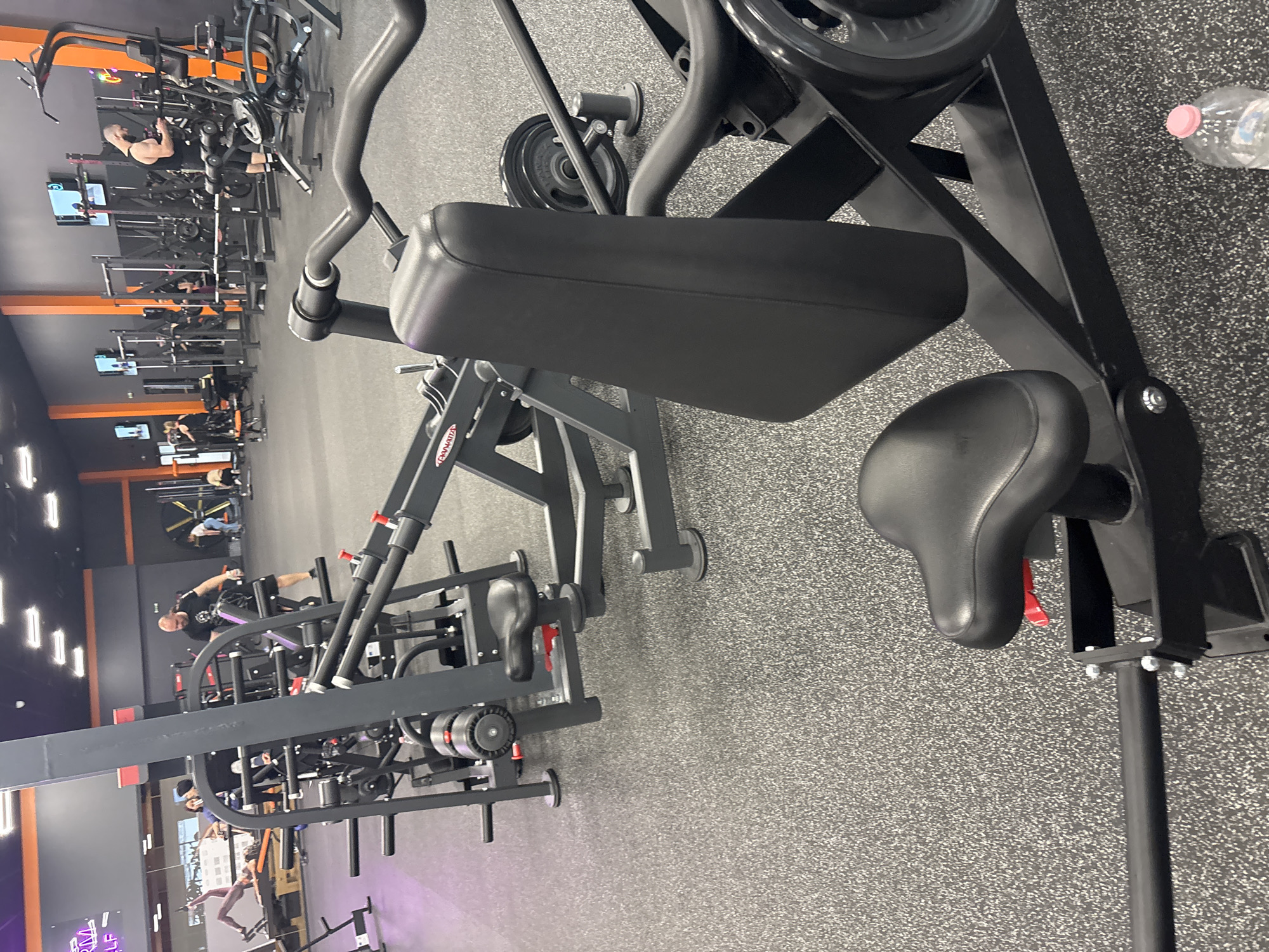 The image, rotated 90 degrees counter-clockwise, captures the interior of a modern gym or fitness center in Trilistnik, Bulgaria. The flooring consists of a grey speckled rubber mat, while the walls are a mix of dark grey/black and bright orange panels. Bright artificial overhead lights illuminate the space.
In the immediate foreground, a large, black-framed piece of gym equipment, likely an upper body machine, is prominent. It features black padded rests, including a rectangular pad and a bicycle-style seat with a visible red adjustment lever. A clear plastic water bottle with a pink cap sits on the floor nearby.
The background showcases numerous other pieces of gym equipment, including various weight machines, benches, and racks, maintaining a consistent aesthetic with black frames and some orange details. Several monitors are mounted on poles throughout the facility. The brand name "PANATTA" is visible on one piece of equipment in the mid-ground. Faintly, outdoor scenery, possibly buildings or trees, can be seen through a distant opening. A purple-lit sign in the lower left background displays partial text, including "RM SELF."
Several people are visible throughout the gym, engaged in exercise. A man with a bald head and beard, wearing a dark tank top, is actively using a weight machine in the middle ground, appearing to perform a seated row. Other individuals are dispersed, utilizing different machines, indicating a typical gym environment during an active training session.