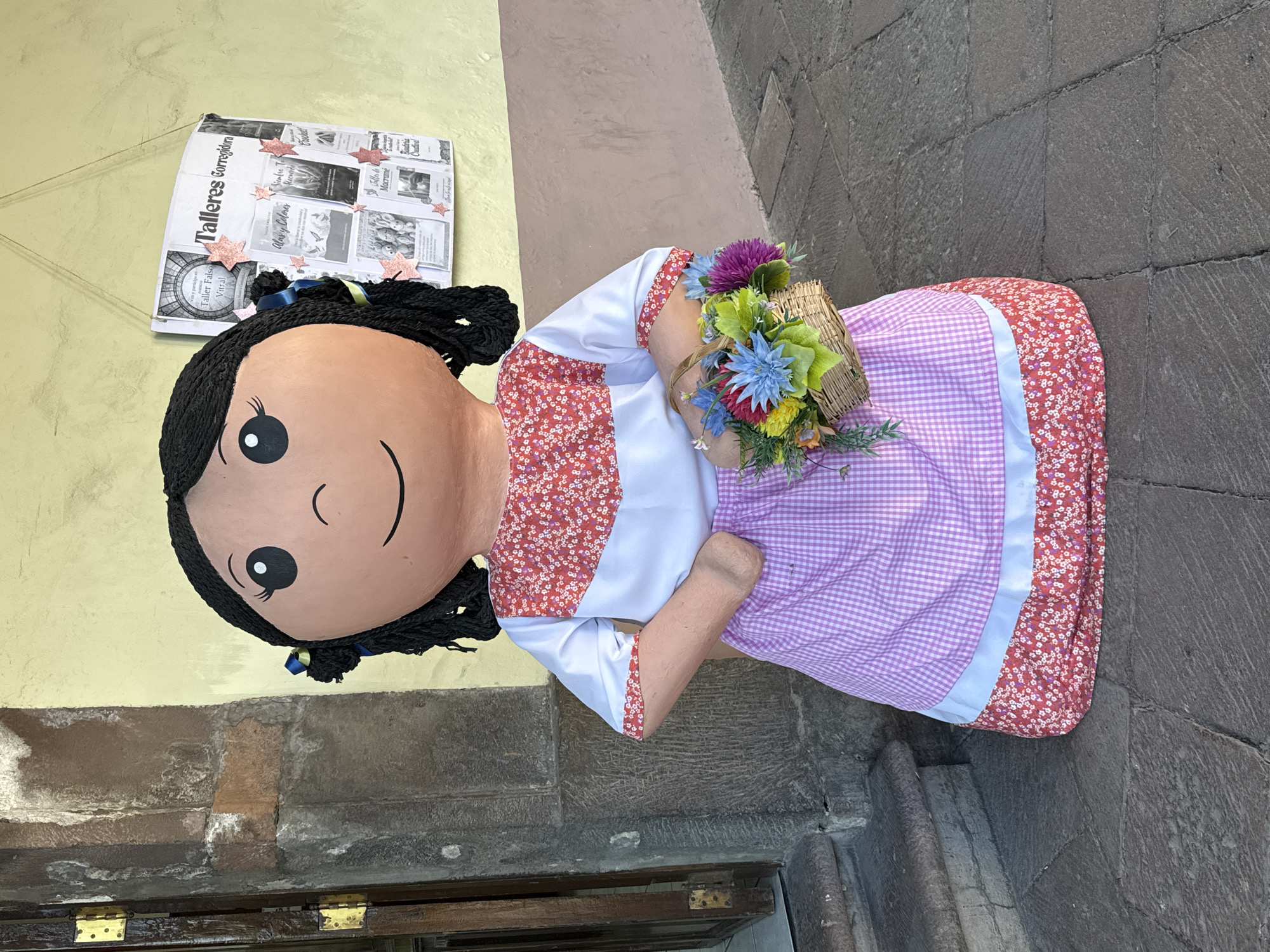 A life-size rag doll figure is displayed outside a building on a stone-paved area. The doll has a large, round face with painted-on features: black eyes, eyelashes, and a simple curved smile. Its black yarn hair is styled into braids with blue ribbons. The doll is dressed in a white blouse with floral print trim, a lavender gingham apron with white trim, and is holding a small basket filled with colorful artificial flowers.
The doll is positioned against a building with a yellow wall. Above the doll's head is a newspaper or pamphlet display advertising "Talleres" (workshops). The background suggests an urban setting, likely a historic area, possibly within the city of Querétaro, Mexico, given the traditional doll design. The doll appears to be leaning against a building's corner, with stone steps visible at the bottom right of the image.