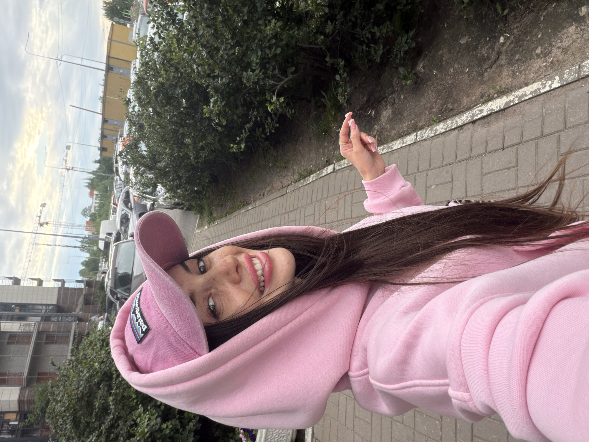In an outdoor, paved urban setting, a young woman with long, dark brown hair, wearing a pink hoodie and a pink baseball cap, smiles directly at the camera, her tongue slightly extended. She is standing on a sidewalk next to a patch of dense green bushes. Behind her, a line of parked cars is visible, and in the distance, a tall building and construction cranes suggest an urban environment. The lighting indicates it is likely late afternoon or early evening. The woman holds a small white object, possibly a flower petal, between her thumb and index finger, with a ring on her right hand. The overall scene appears to be a selfie taken in an ordinary city location in Russia.